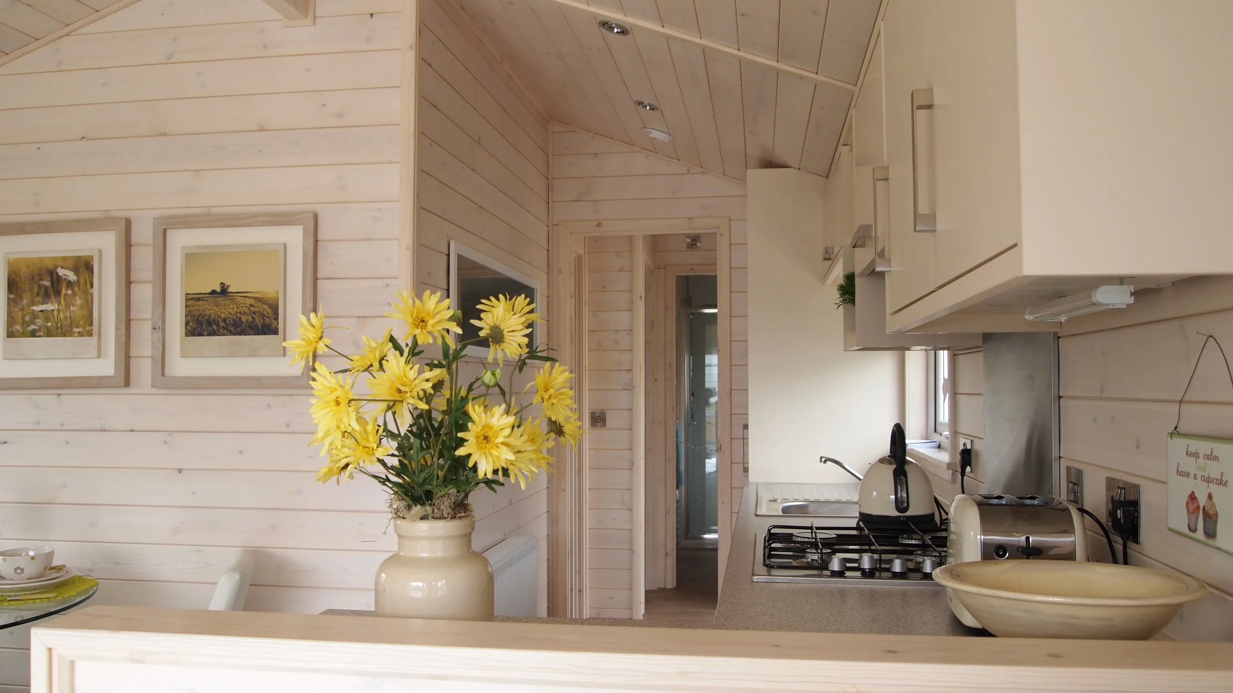 A kitchen with light wood paneling, white cabinets, a stove with a tea kettle, a kitchen faucet, a small window, a decorative bowl, and a large vase of yellow flowers on the counter.