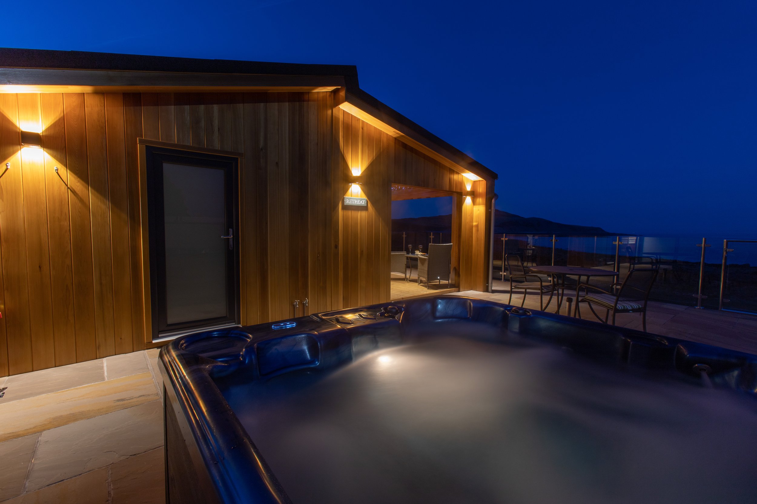 Outdoor hot tub at night on a wooden deck, with a view of the ocean and mountains in the distance. The house has wood paneling and is lit with warm lights.