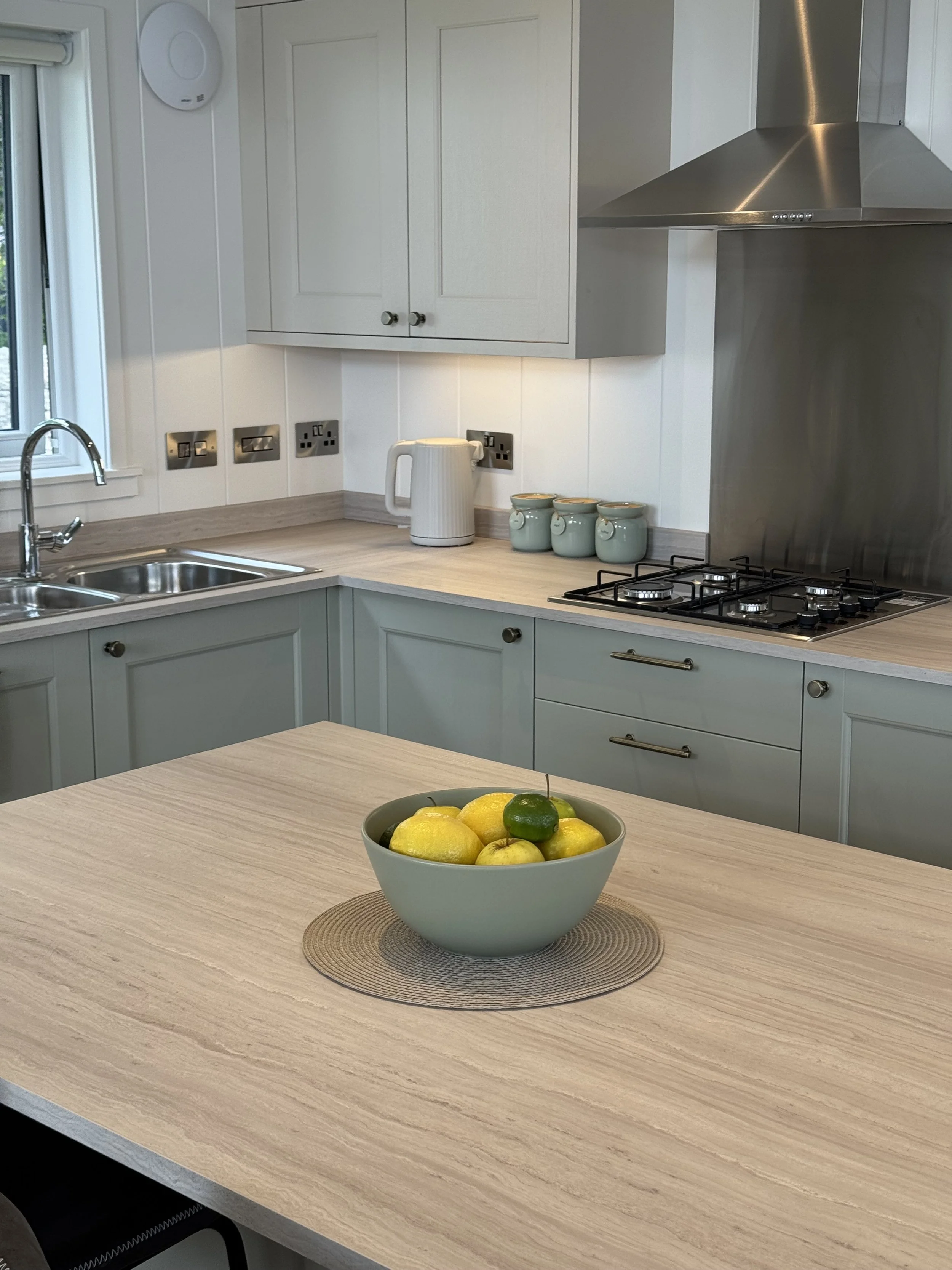 A modern kitchen with light-colored cabinets, a stainless steel range hood, a double sink, and a countertop with a white electric kettle and three small green jars. There is a bowl of lemons and a lime on a round placemat on the kitchen island.