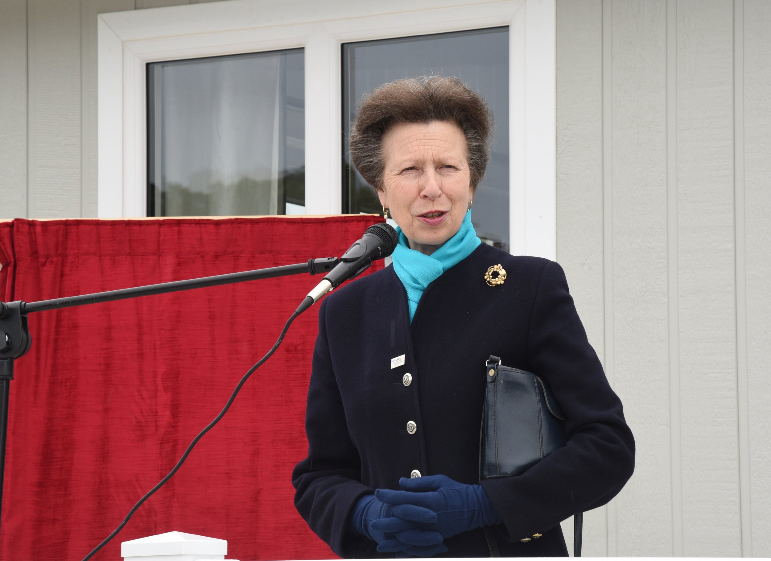 A woman standing in front of a microphone outdoors, wearing a dark coat, blue gloves, and a light blue scarf, holding a black purse. Behind her is a red curtain and a white building with windows.