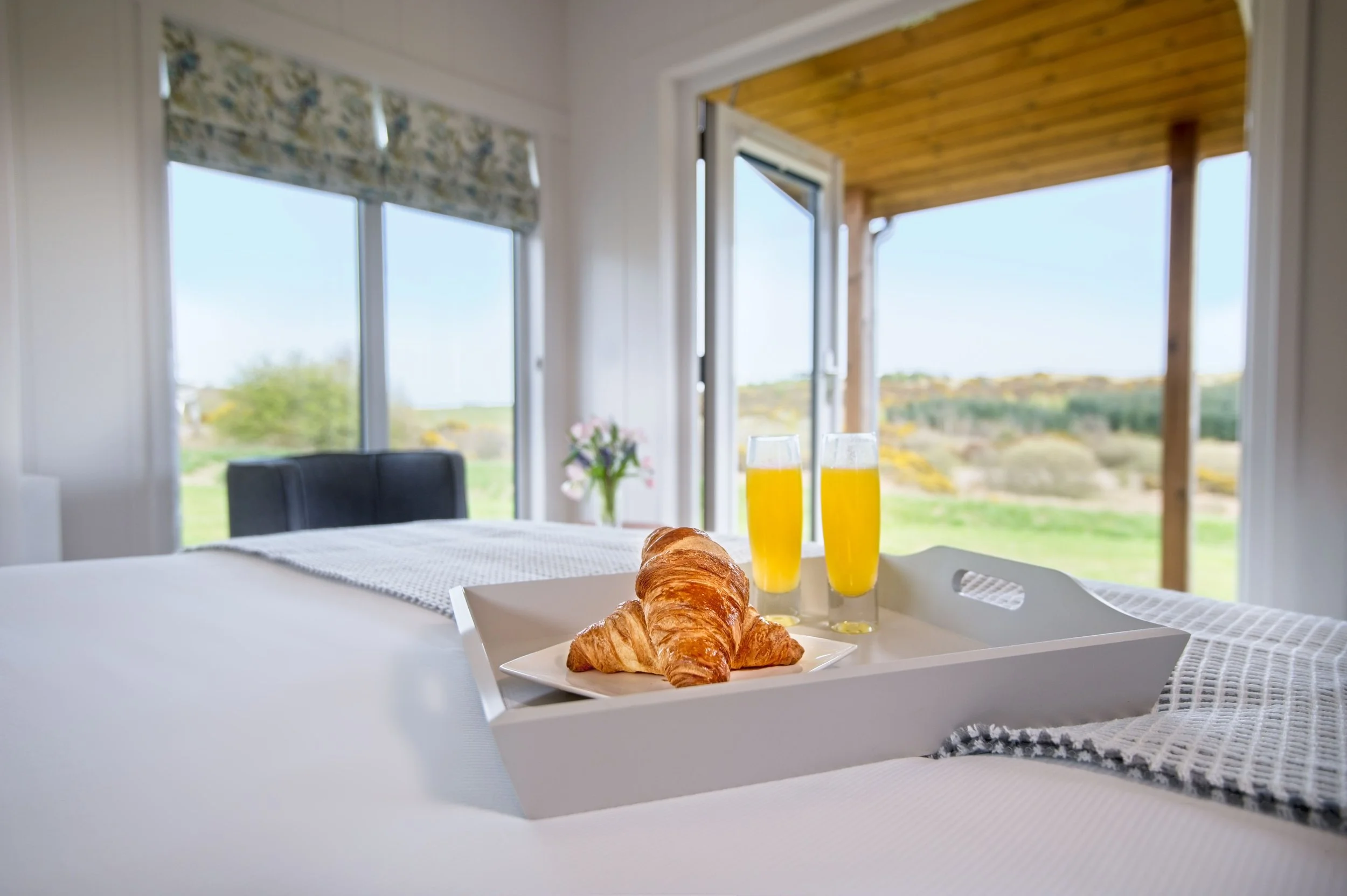 Breakfast tray on a bed with croissants and two glasses of orange juice, in a bedroom with large windows showing a scenic outdoor view.