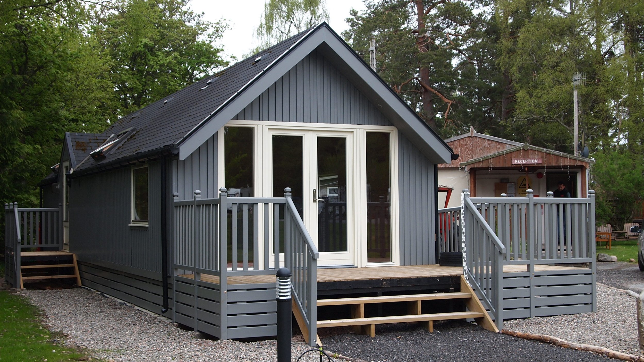 Gray tiny house with front porch and stairs in a forested area, with a reception building in the background.