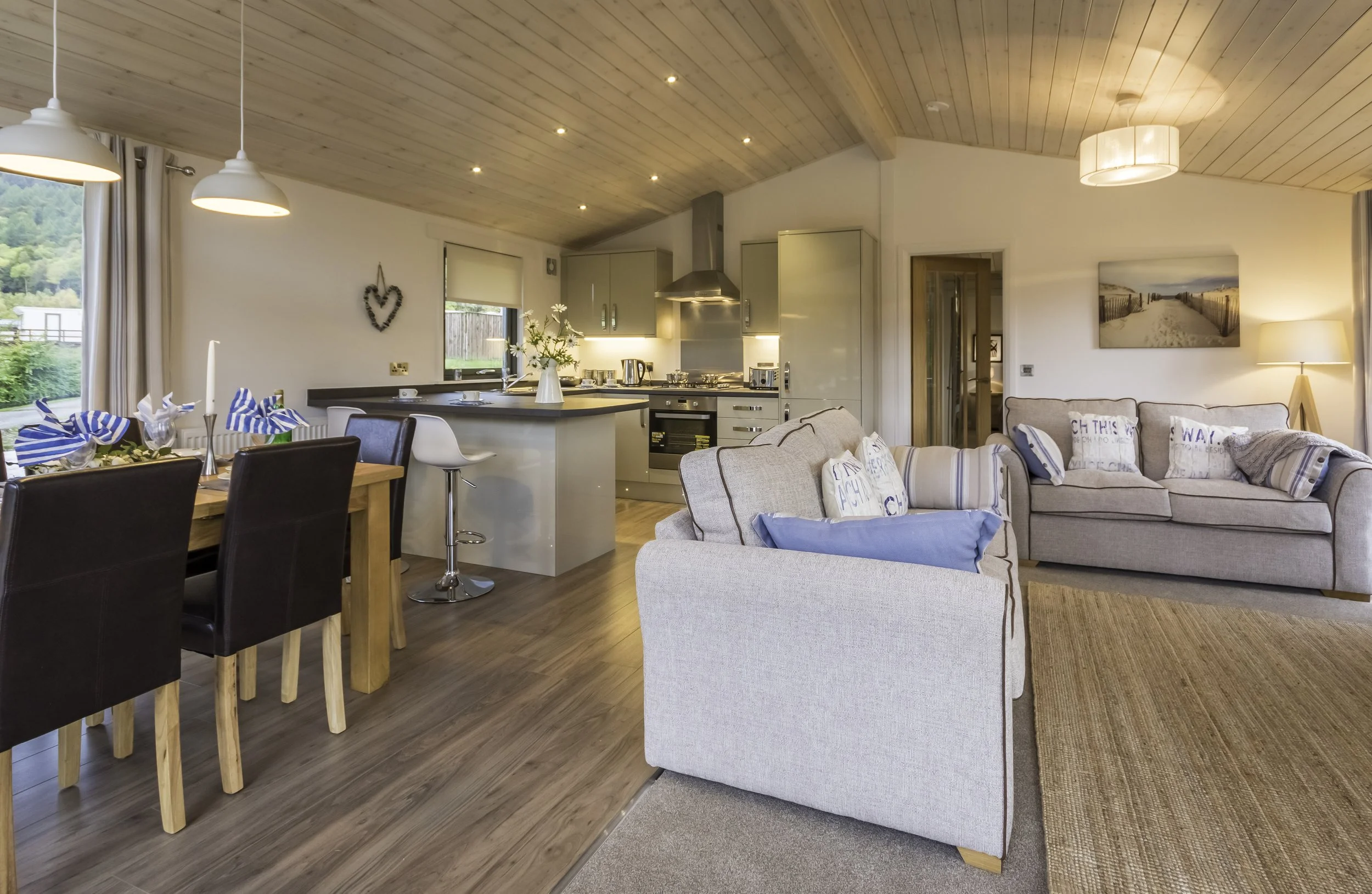 Open-concept living room and kitchen with wooden ceiling, beige sofas, dining table with black chairs, and a view of outdoors through windows.