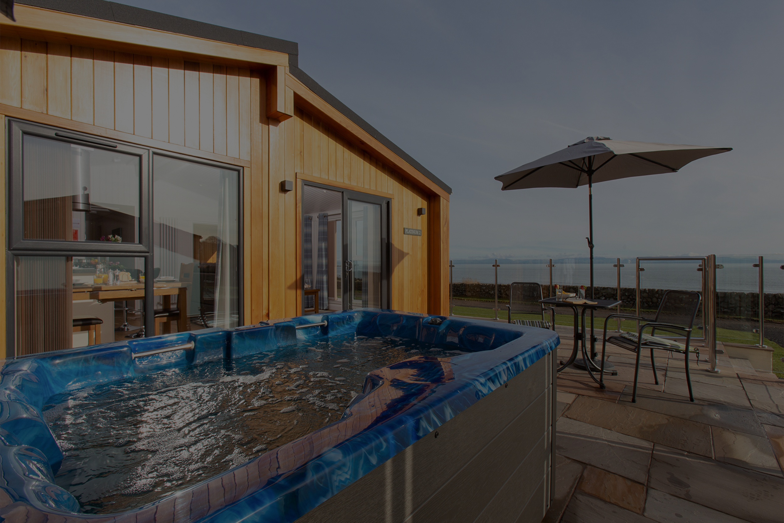 Outdoor patio with hot tub, table, chairs, and umbrella, overlooking body of water