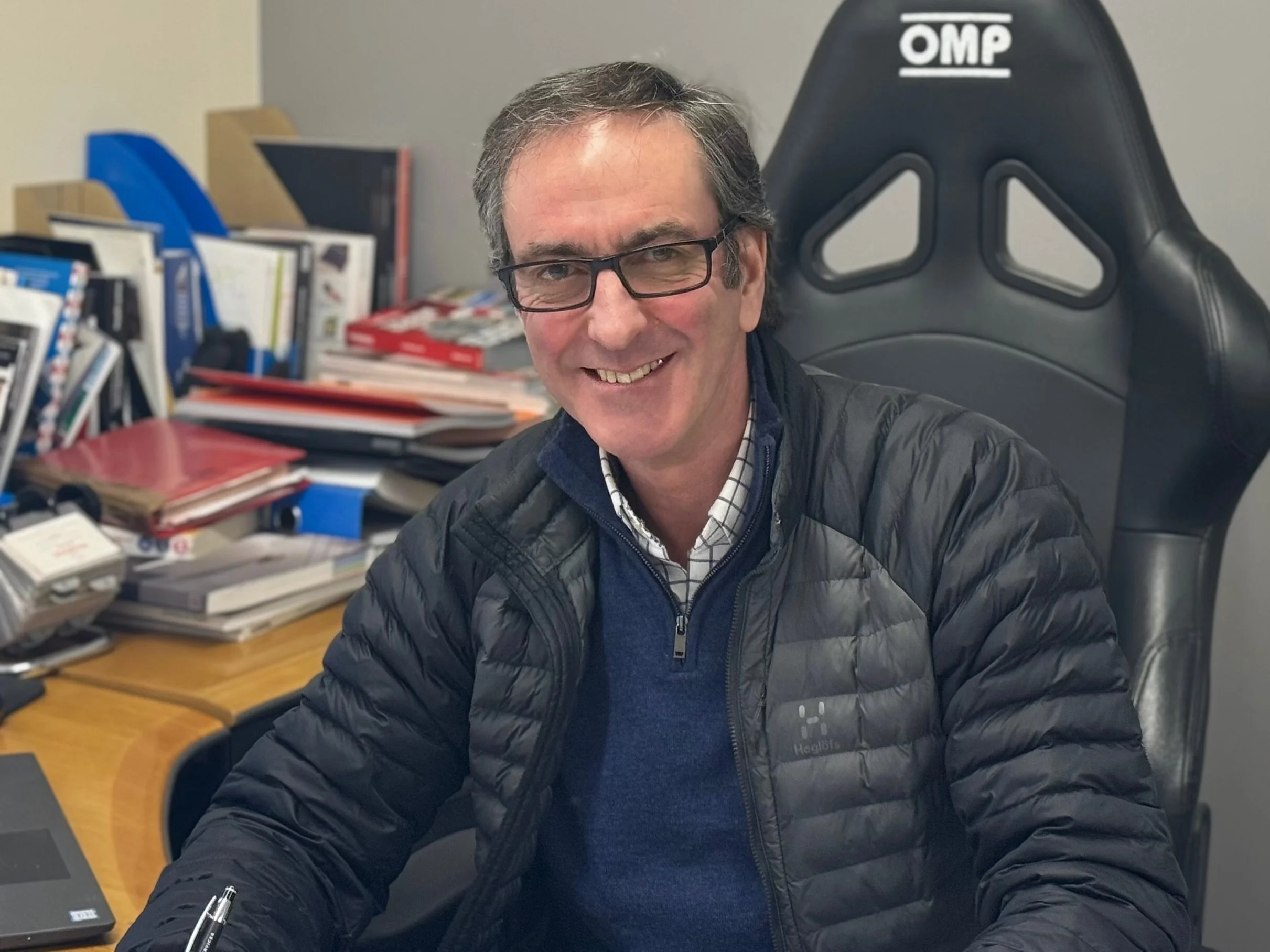 A smiling man wearing glasses and a black puffer jacket sitting at a cluttered desk with books and papers, in front of a black OMP gaming chair.