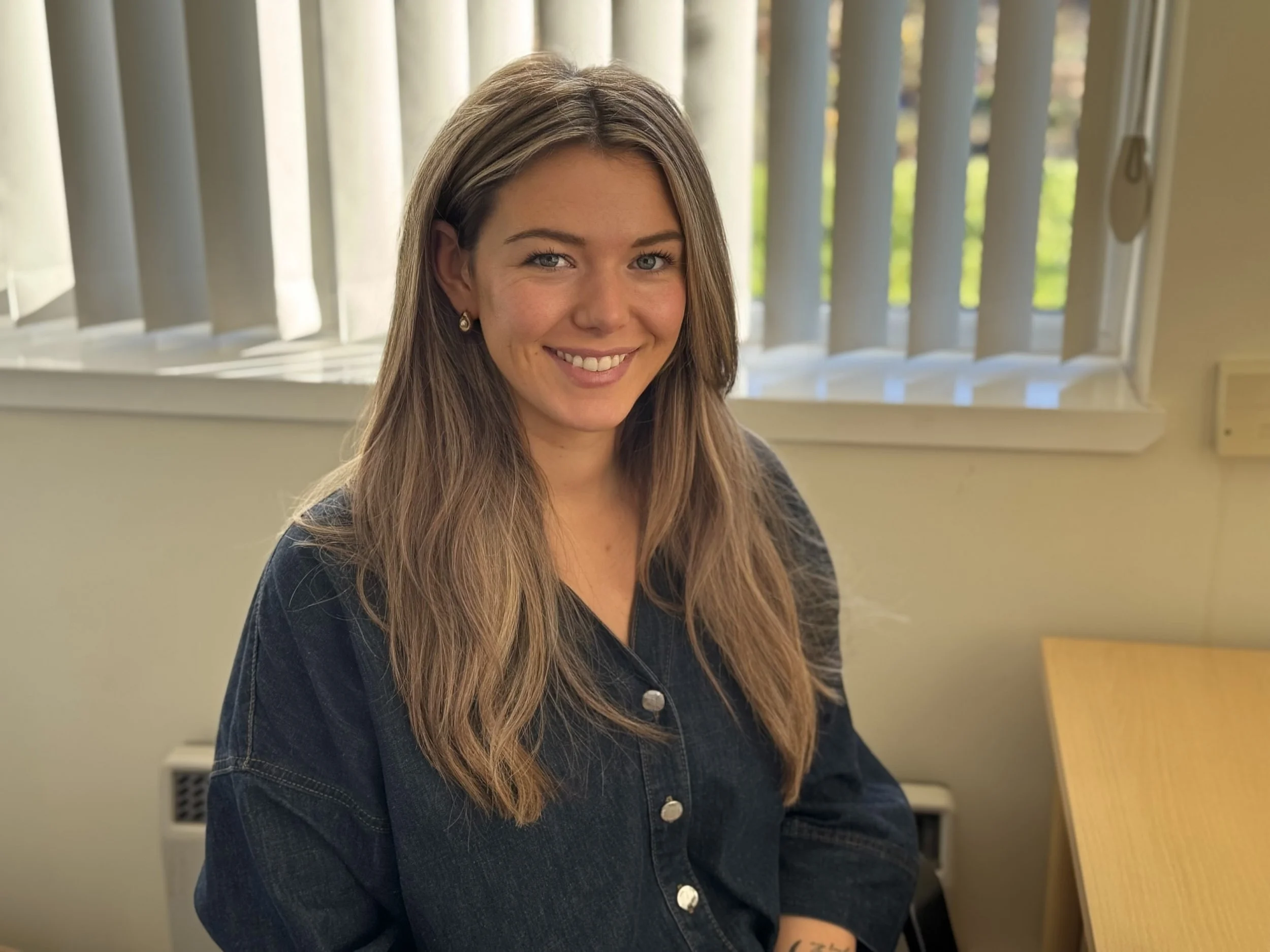 A woman with long brown hair smiling, wearing a dark denim jacket, sitting indoors near a window with vertical blinds and a light-colored wall.
