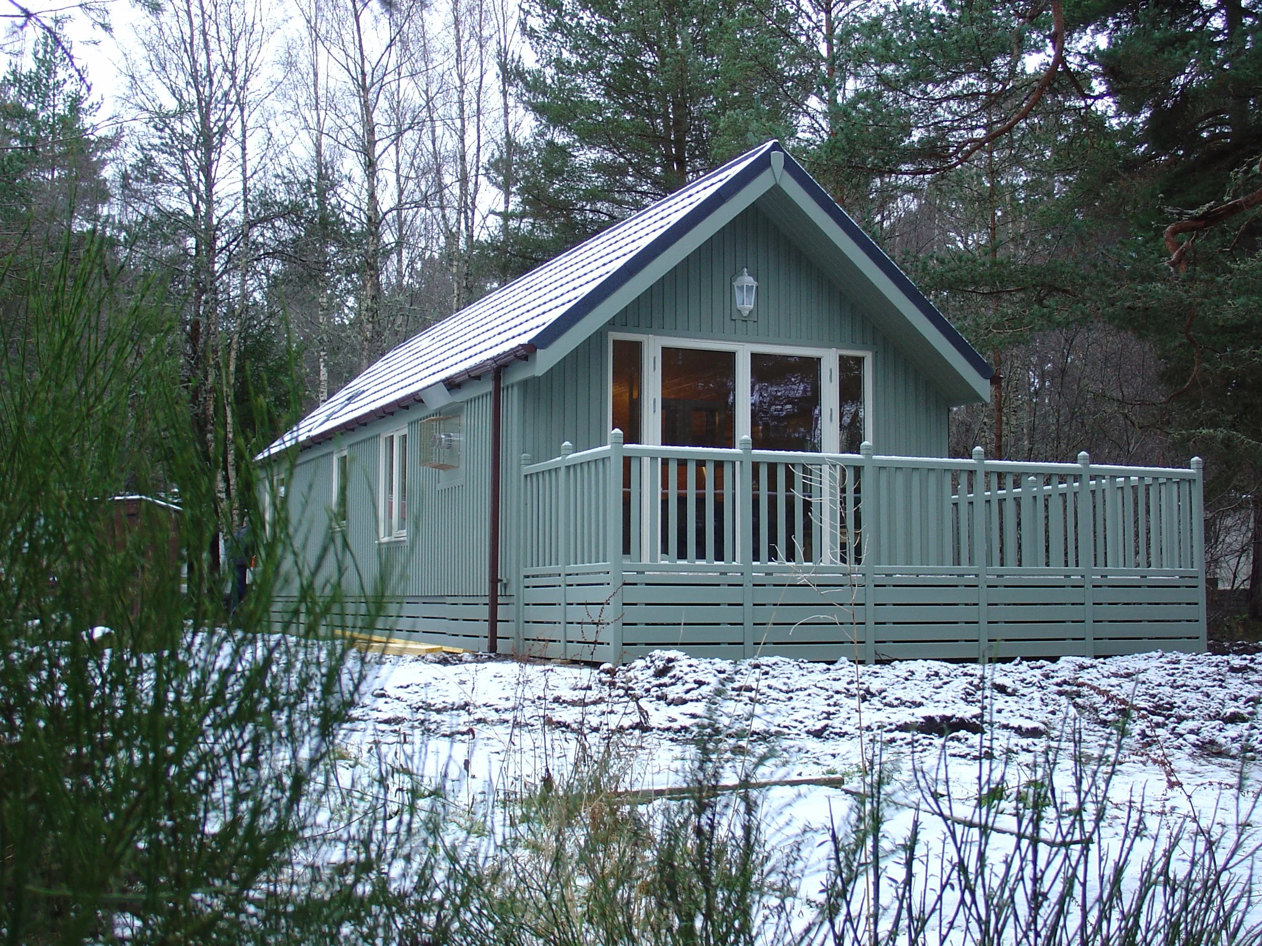 A light blue wooden house with a snow-covered roof, located in a wooded area with trees and snow on the ground.