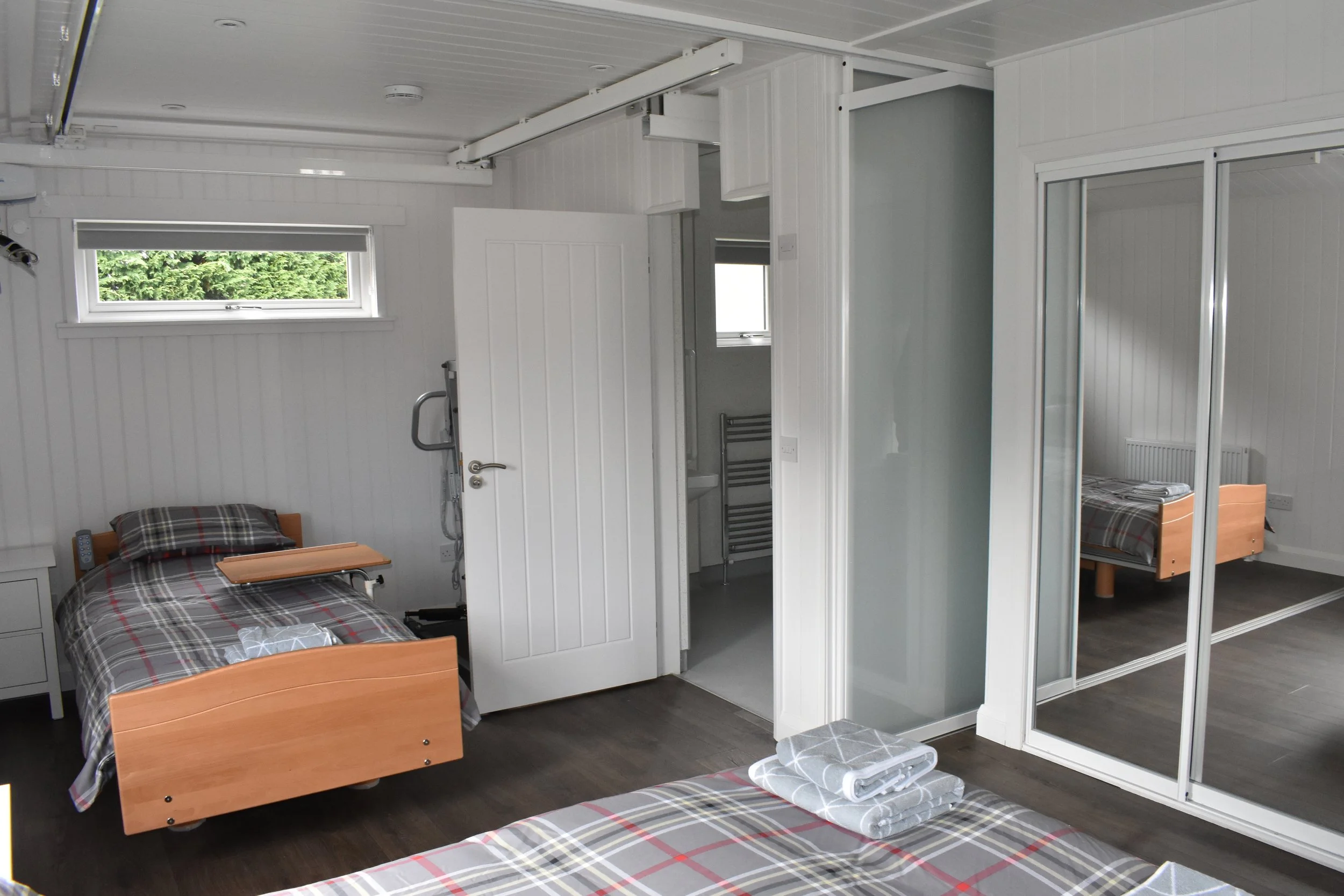 A bedroom with two single beds with plaid bedding, one with a pillow, a mirror sliding door closet, a small window, and a door leading to a small bathroom with a sink and towel rack. Towels are folded on the bed.