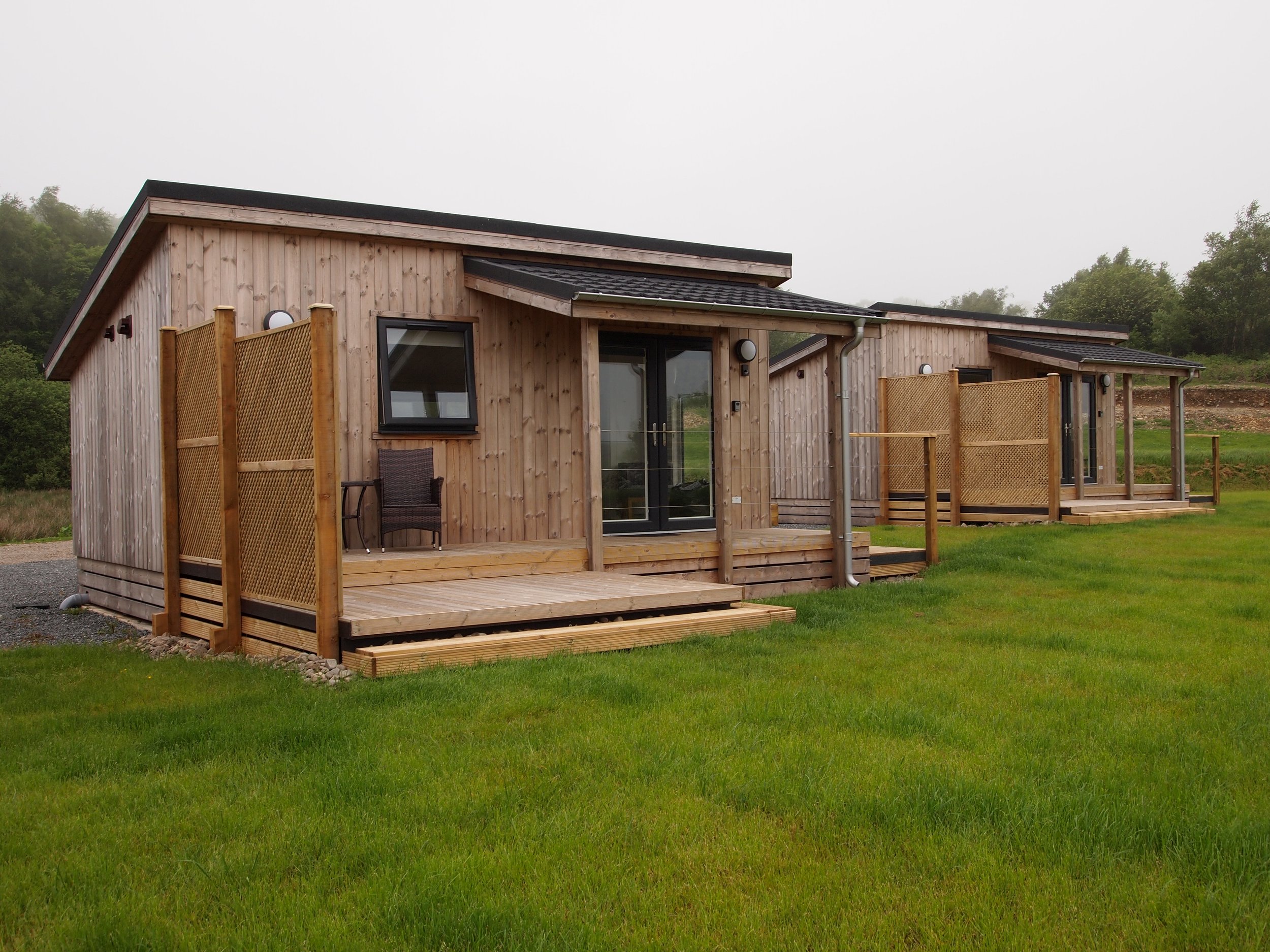 Two modern wooden houses with small decks, set in a grassy landscape with hills and trees in the background, under a cloudy sky.