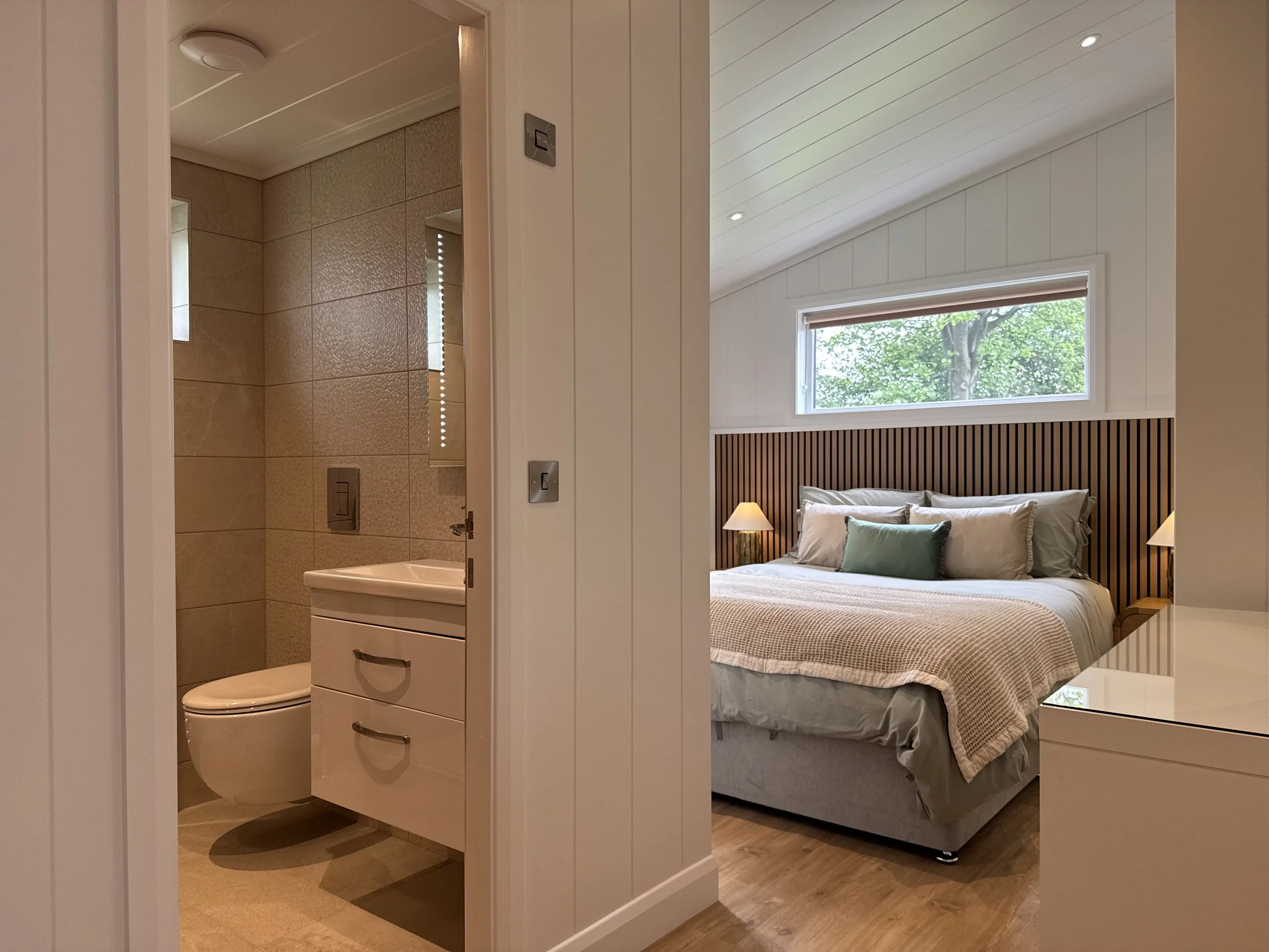 A bedroom with a large bed with a wooden headboard, pillows, and a beige blanket, and a window showing green trees outside. An adjacent bathroom with beige tiled walls, a toilet, a small sink, and small vertical windows.
