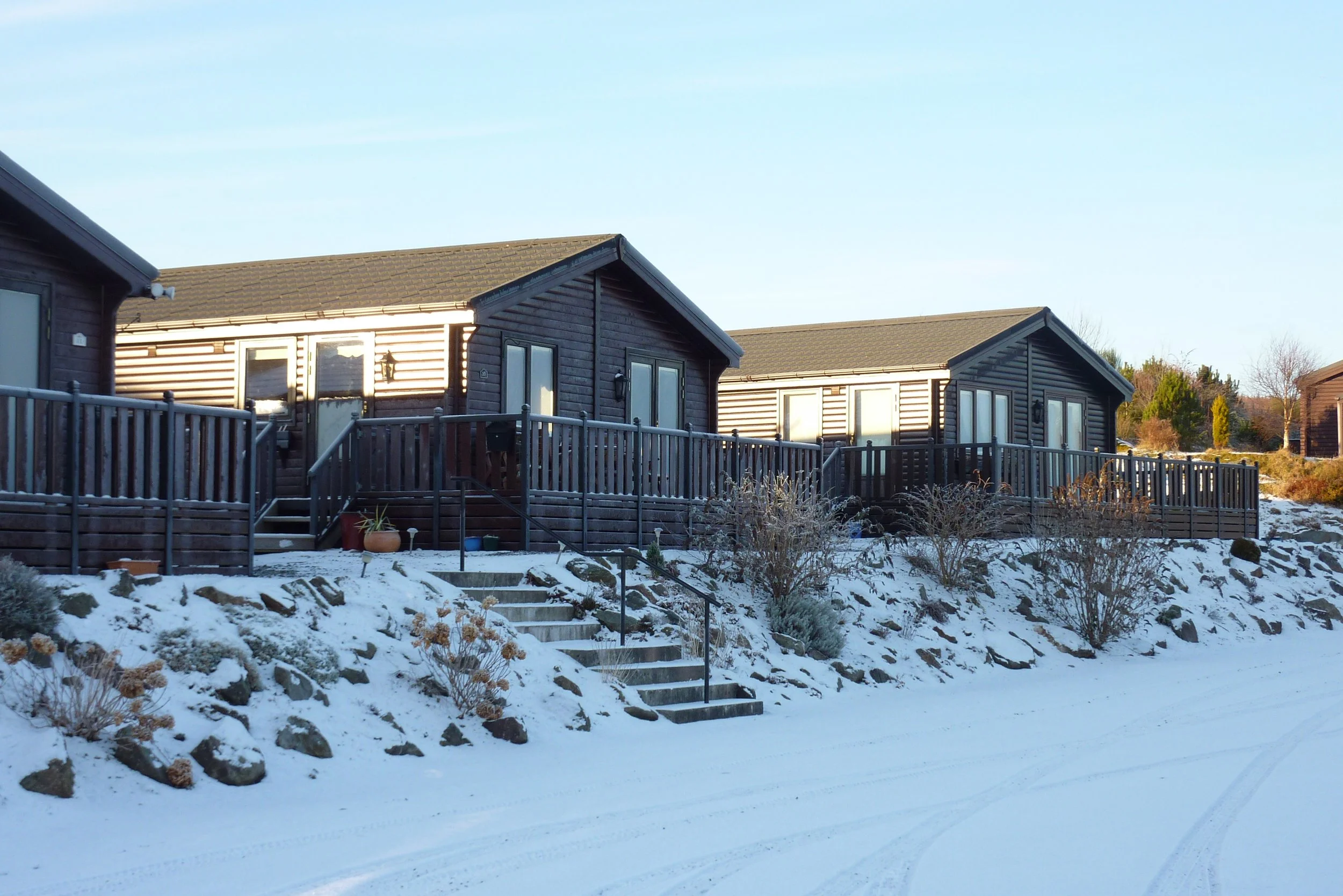 Three dark-colored houses with decks on snowy ground under a clear blue sky.