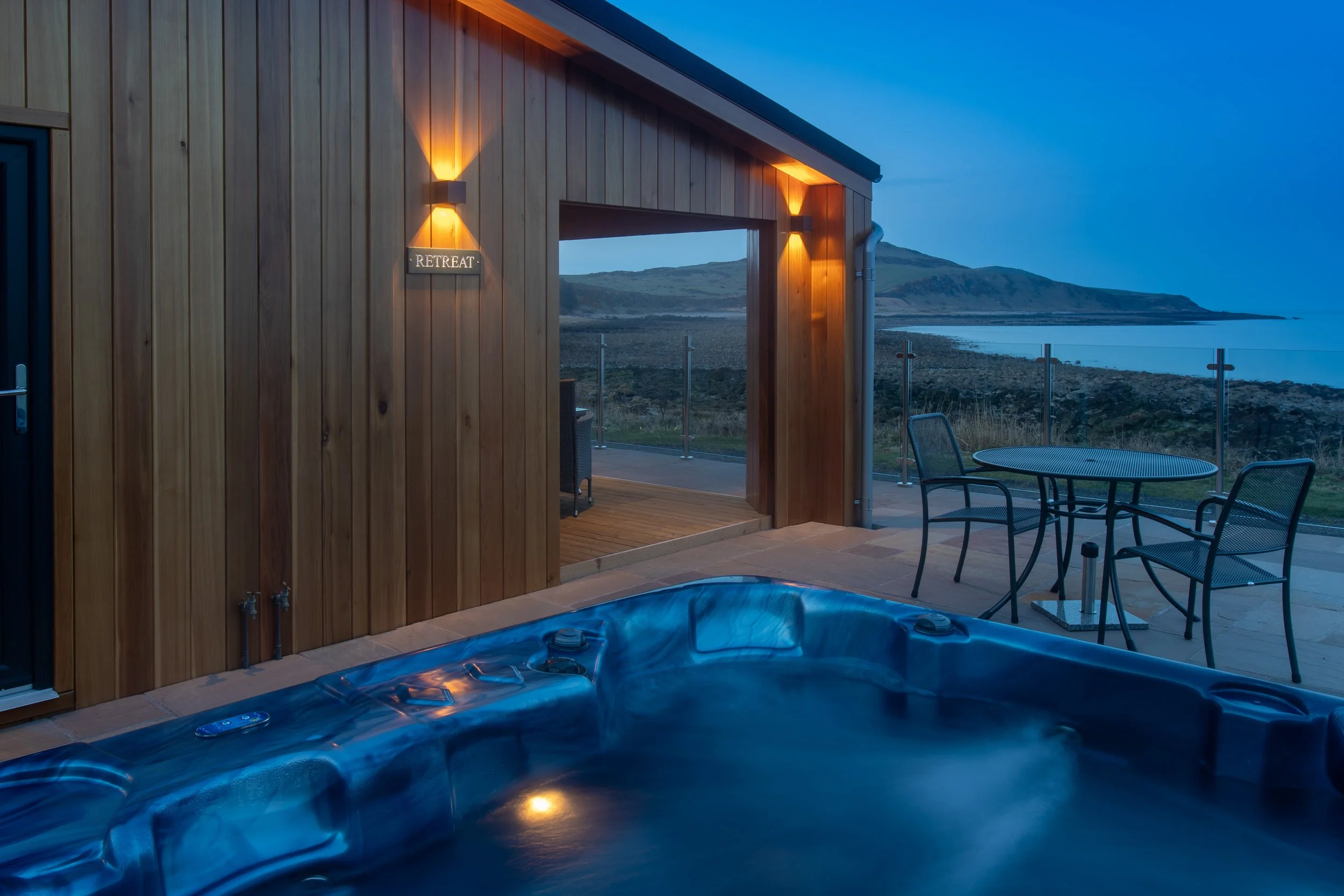 Outdoor hot tub on a patio overlooking water and hills at dusk, with a small table and chairs, wooden building with lights, and a sign that says 'RETREAT'.