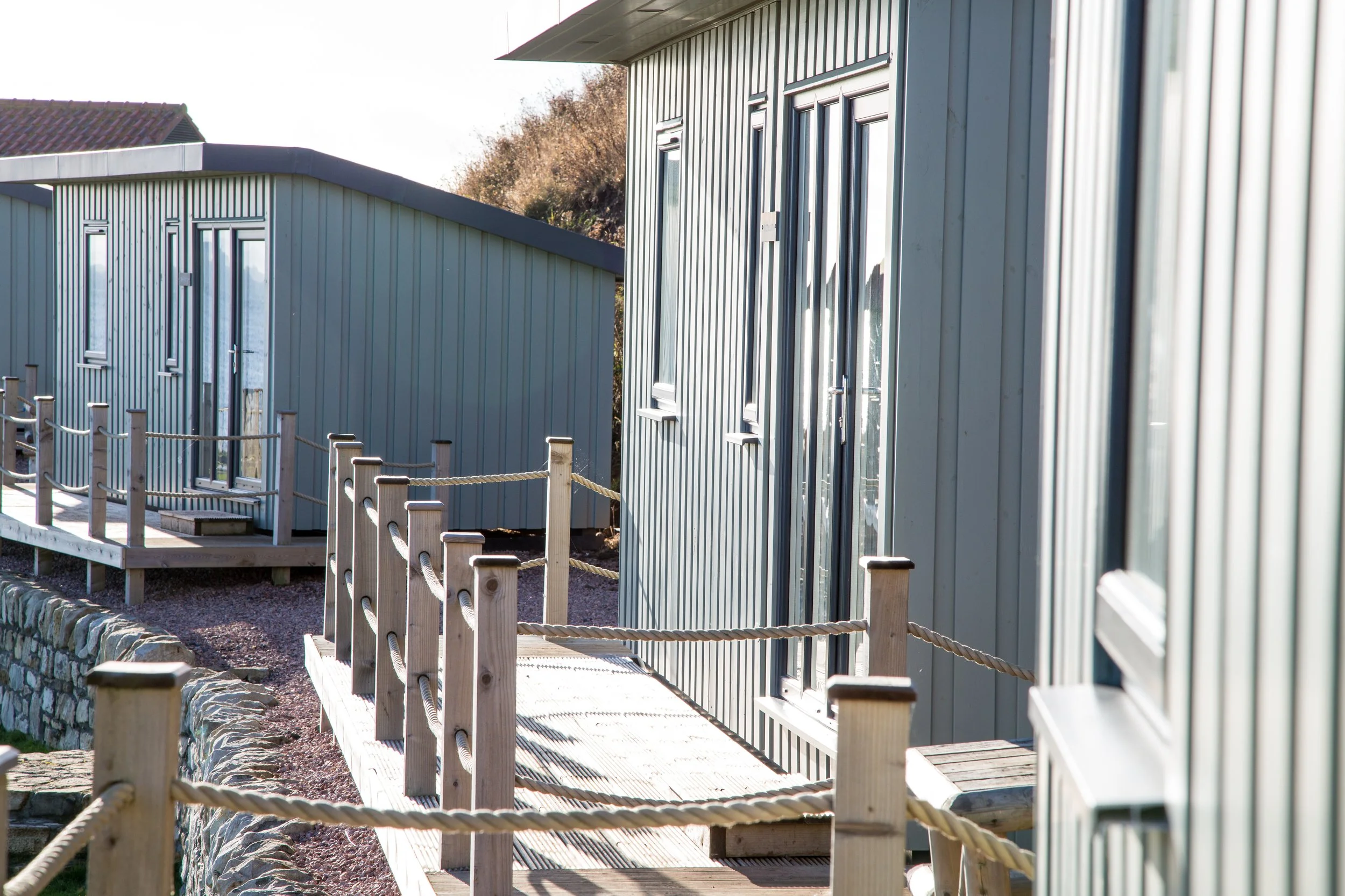 Multiple modern gray metal buildings with large windows, connected by wooden walkways with rope railings, in a sunny outdoor setting.