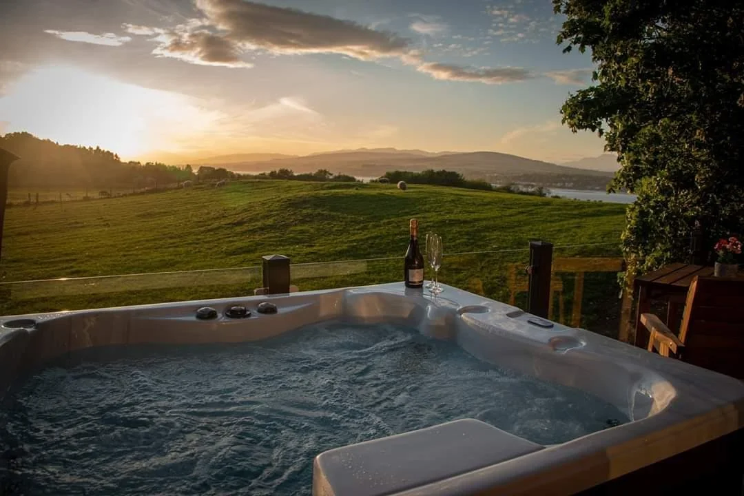 Hot tub on a deck overlooking a green field and hills in the distance during sunset, with a bottle of wine and two glasses on the edge.