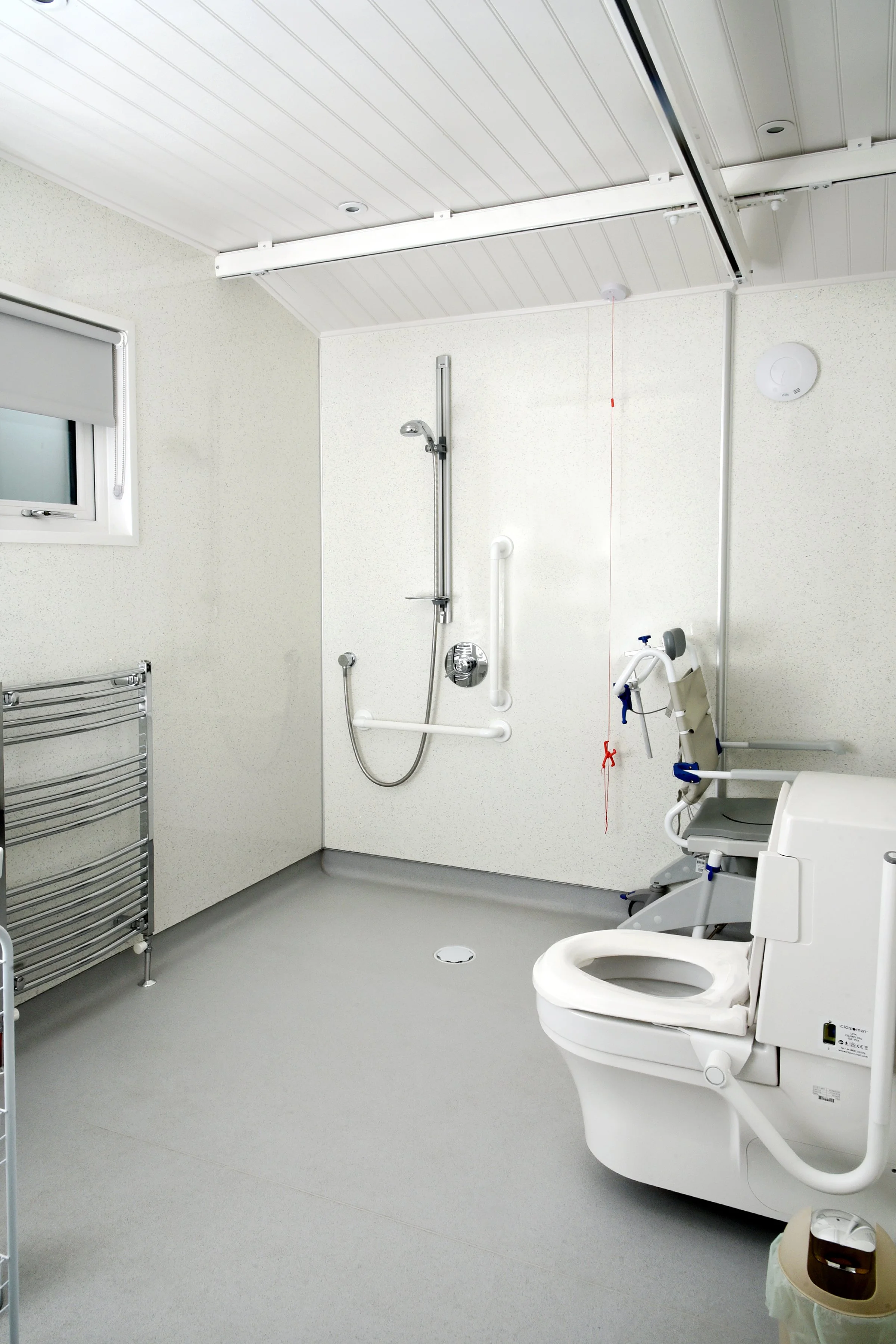 A clean hospital bathroom with a toilet, grab bars, and a shower area with a handheld showerhead, wall-mounted controls, and a ceiling rail for assistance, featuring a small window and minimal decor.