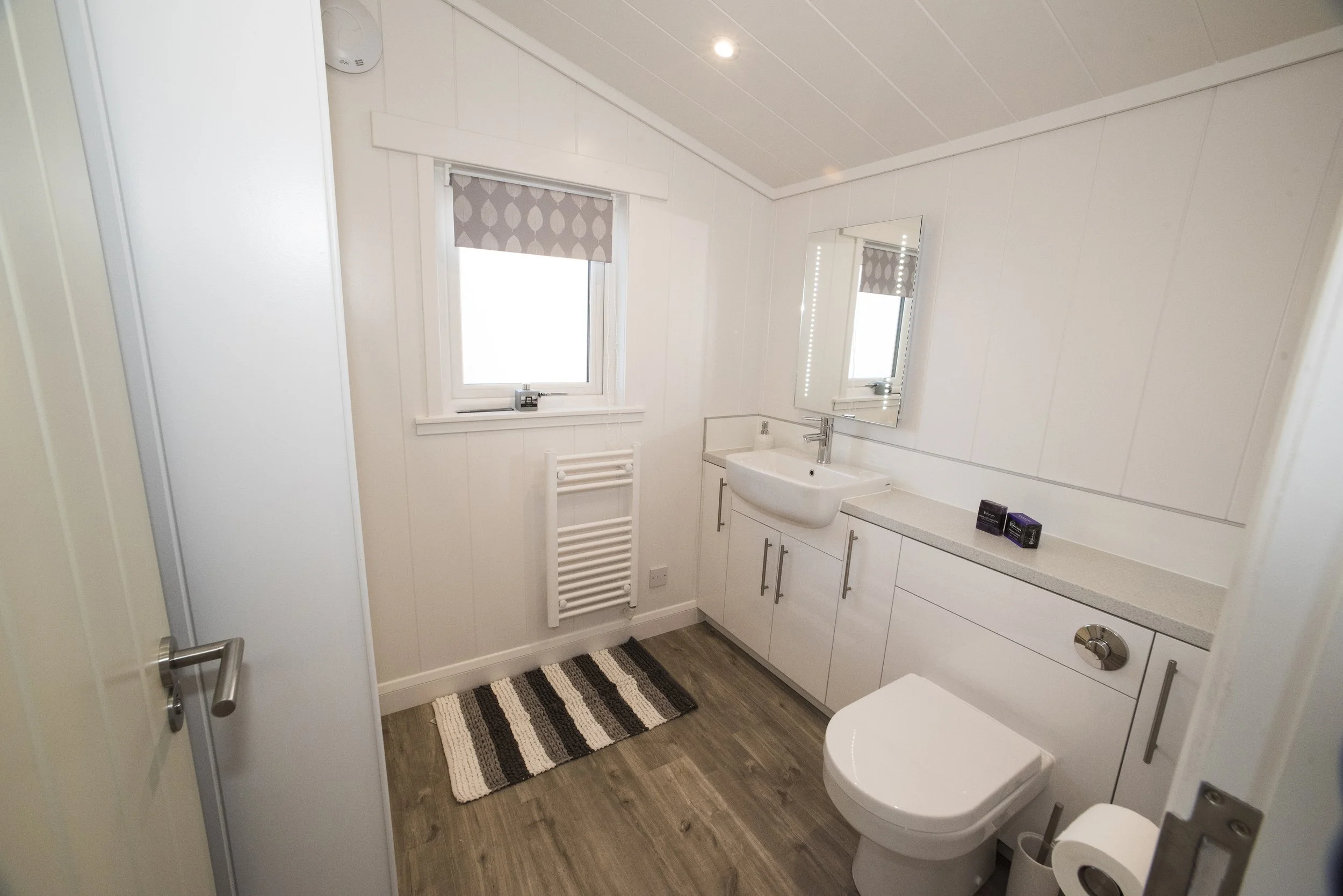 A small, modern bathroom with white walls, a window with a gray patterned blind, a white vanity sink with a large mirror above, a white toilet, a striped rug on wood-look flooring, and a radiator heater.