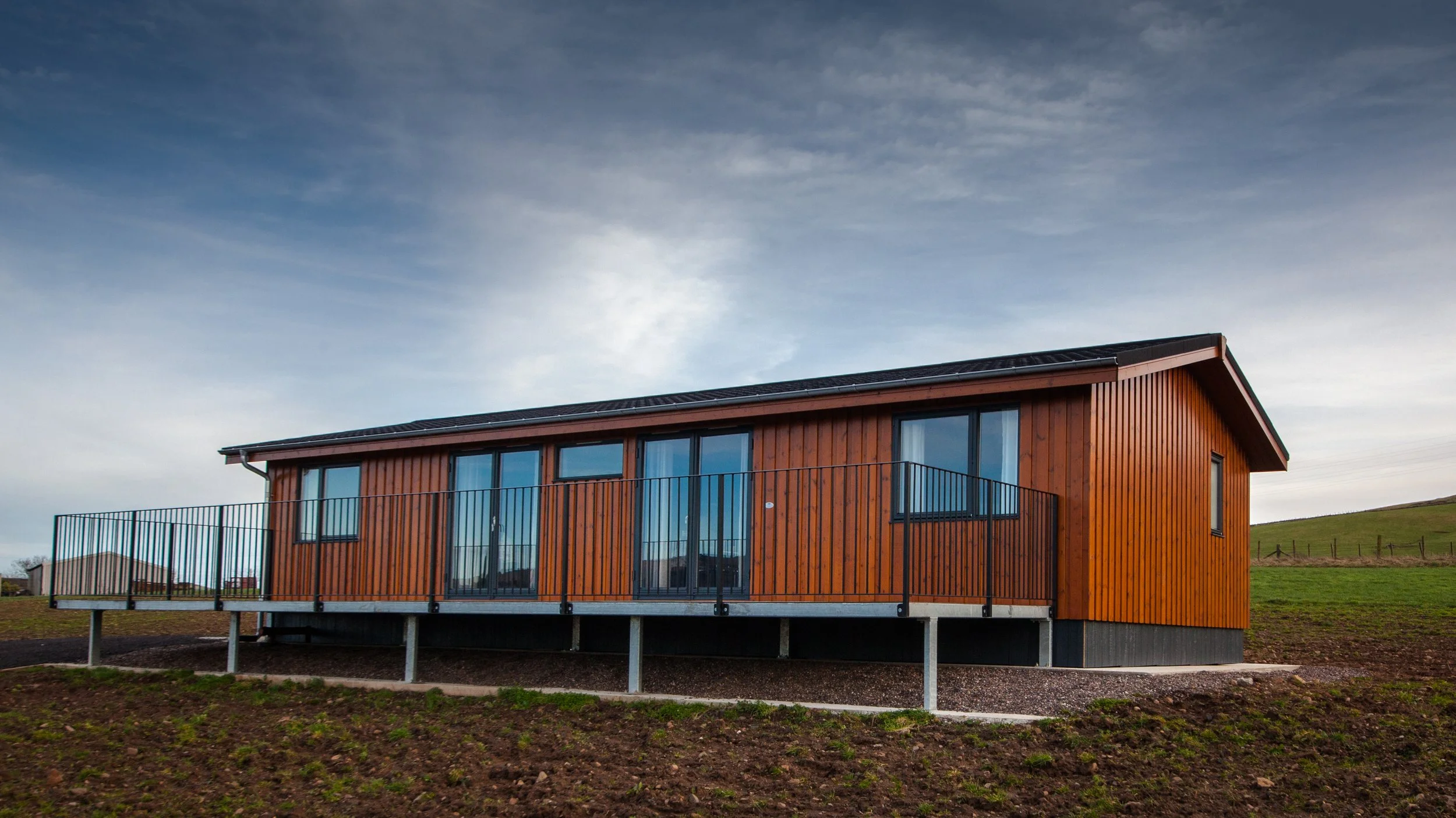 A modern wooden house with large glass windows and a metal railing balcony, elevated on stilts in a rural area with green hills and a cloudy sky.