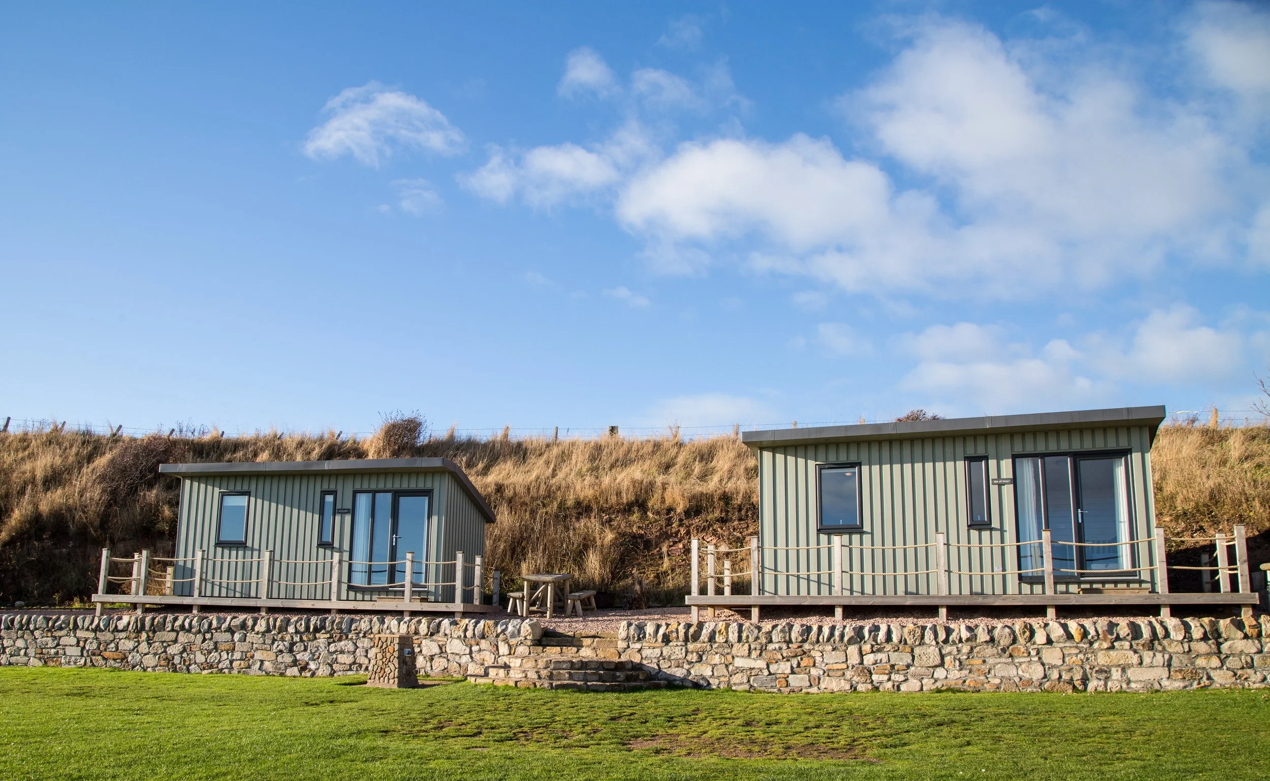 Two modern small houses with grey siding and glass doors, set on a stone and wooden terrace overlooking grassy area, under a partly cloudy blue sky.