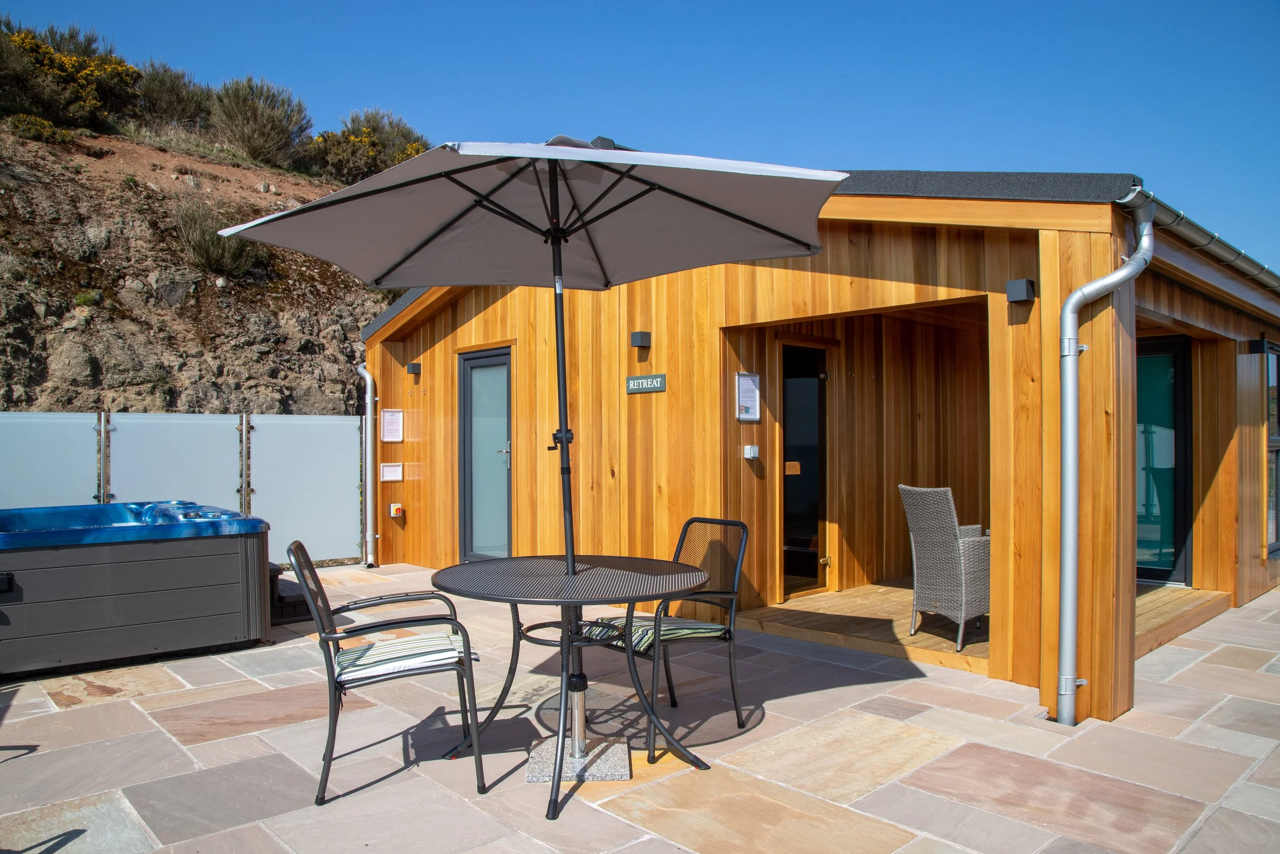 Outdoor patio with a black round table, three metal chairs, a large umbrella, hot tub, and wooden building labeled 'RETREAT' against a rocky hillside and clear blue sky.