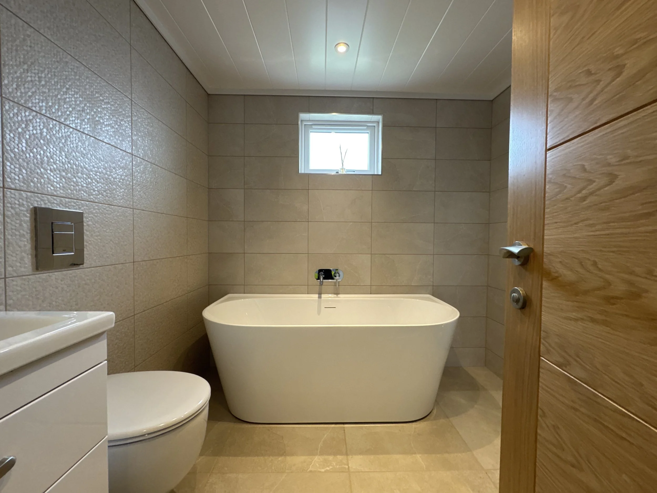 Modern bathroom with beige tiled walls and floor, a white bathtub under a small window, a white toilet, and a wooden door partially open.
