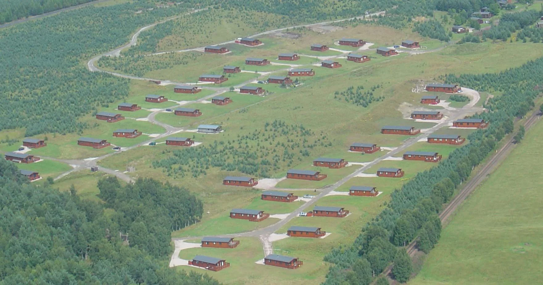 Aerial view of a hillside residential area with multiple single-story wooden houses, winding roads, and surrounding green fields and trees.