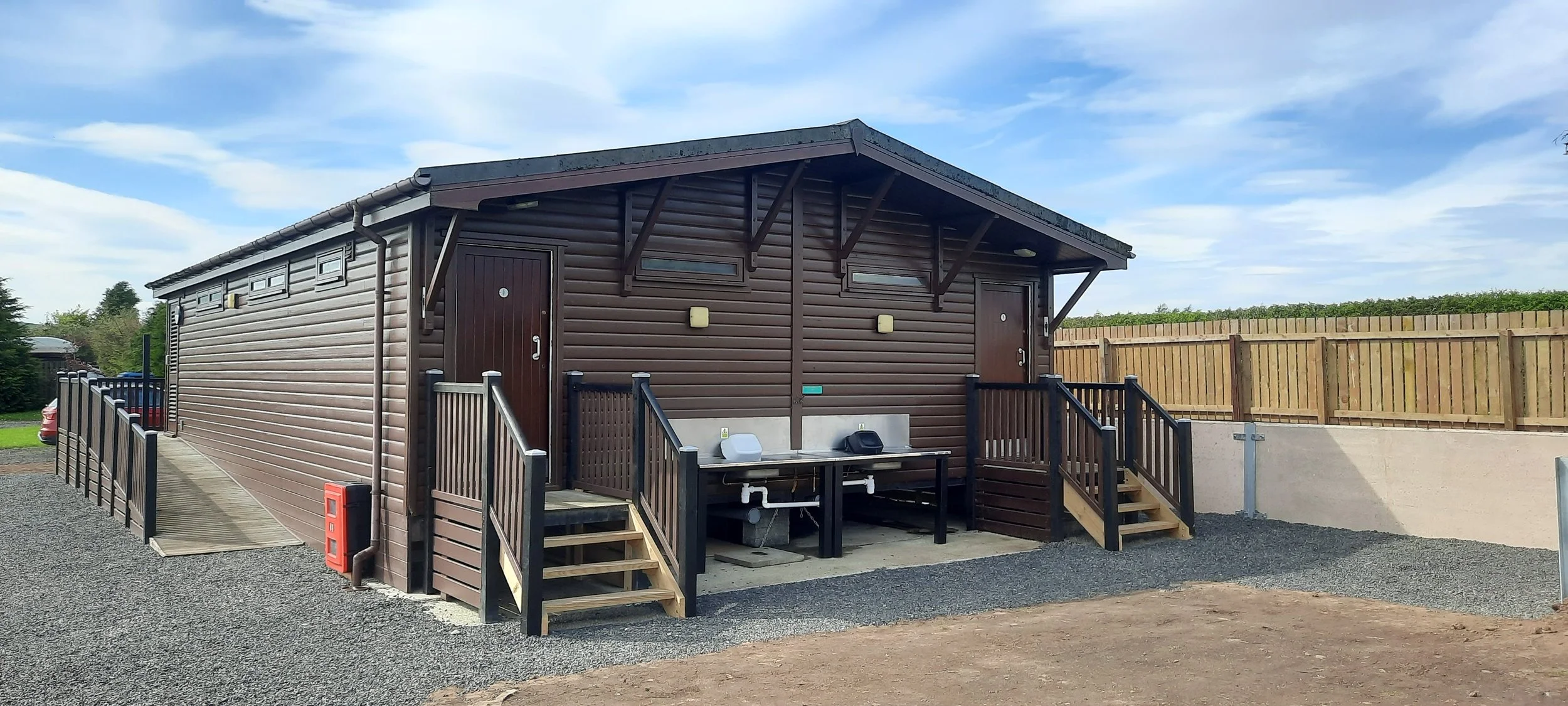 Small brown wooden building with two doors, small stairs, outdoor sinks, and a fenced area, set against a partly cloudy sky.