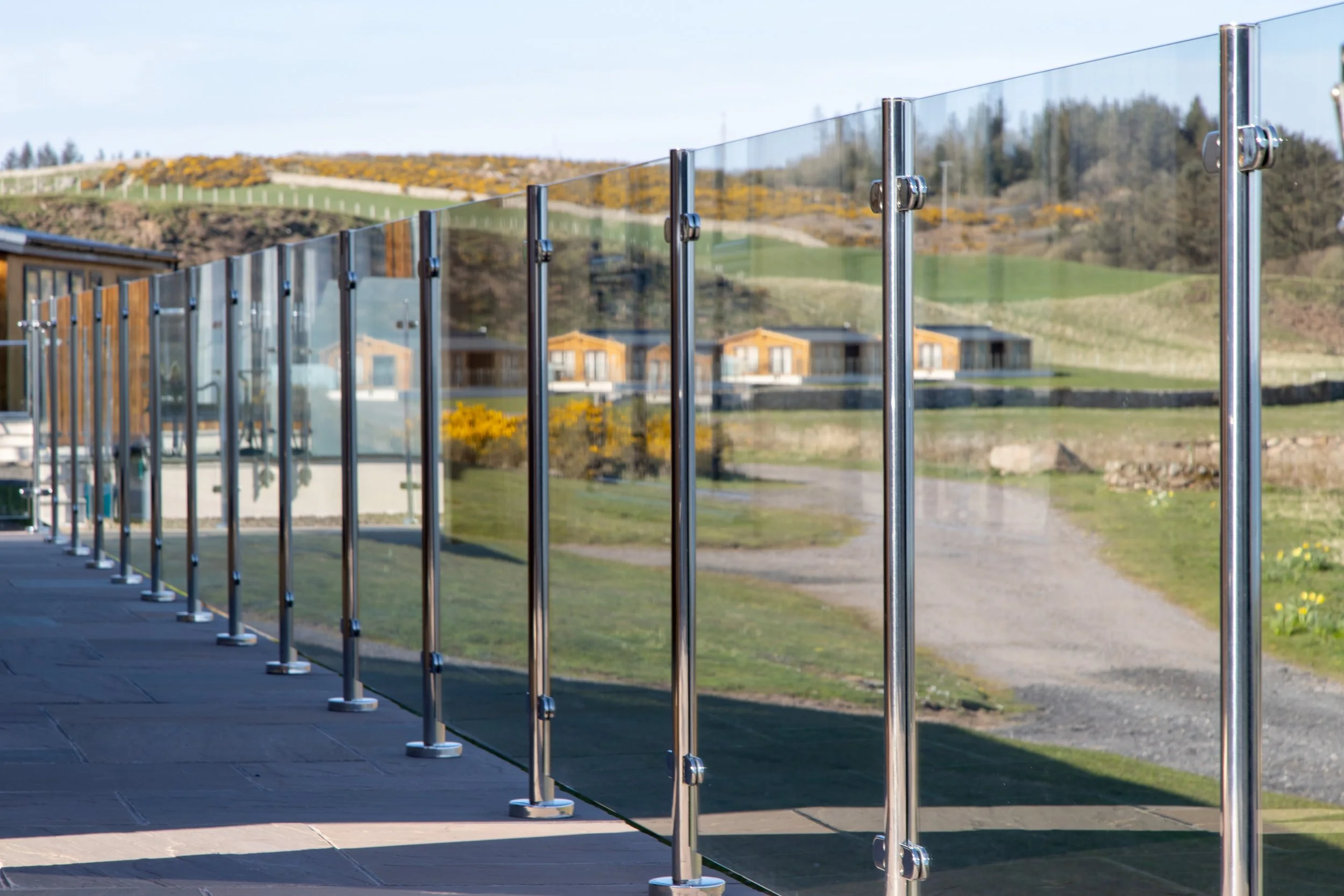 Glass balcony railing with metal posts overlooking a grassy landscape with hills, trees, and houses in the background.