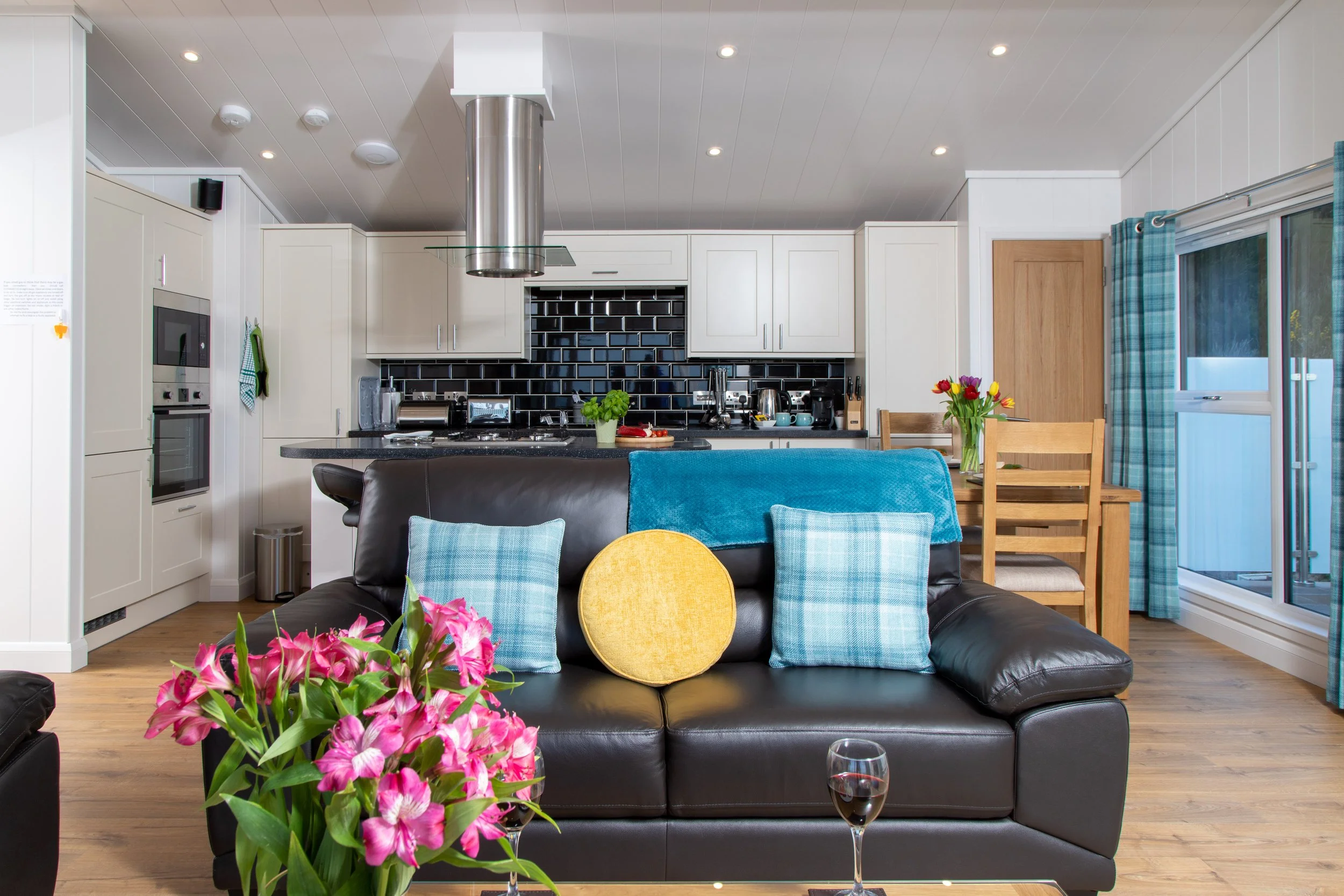Open-concept living room and kitchen with black leather sofa, colorful pillows, a glass of red wine, a vase of pink flowers, wooden table with chairs, and white cabinetry with black tile backsplash.