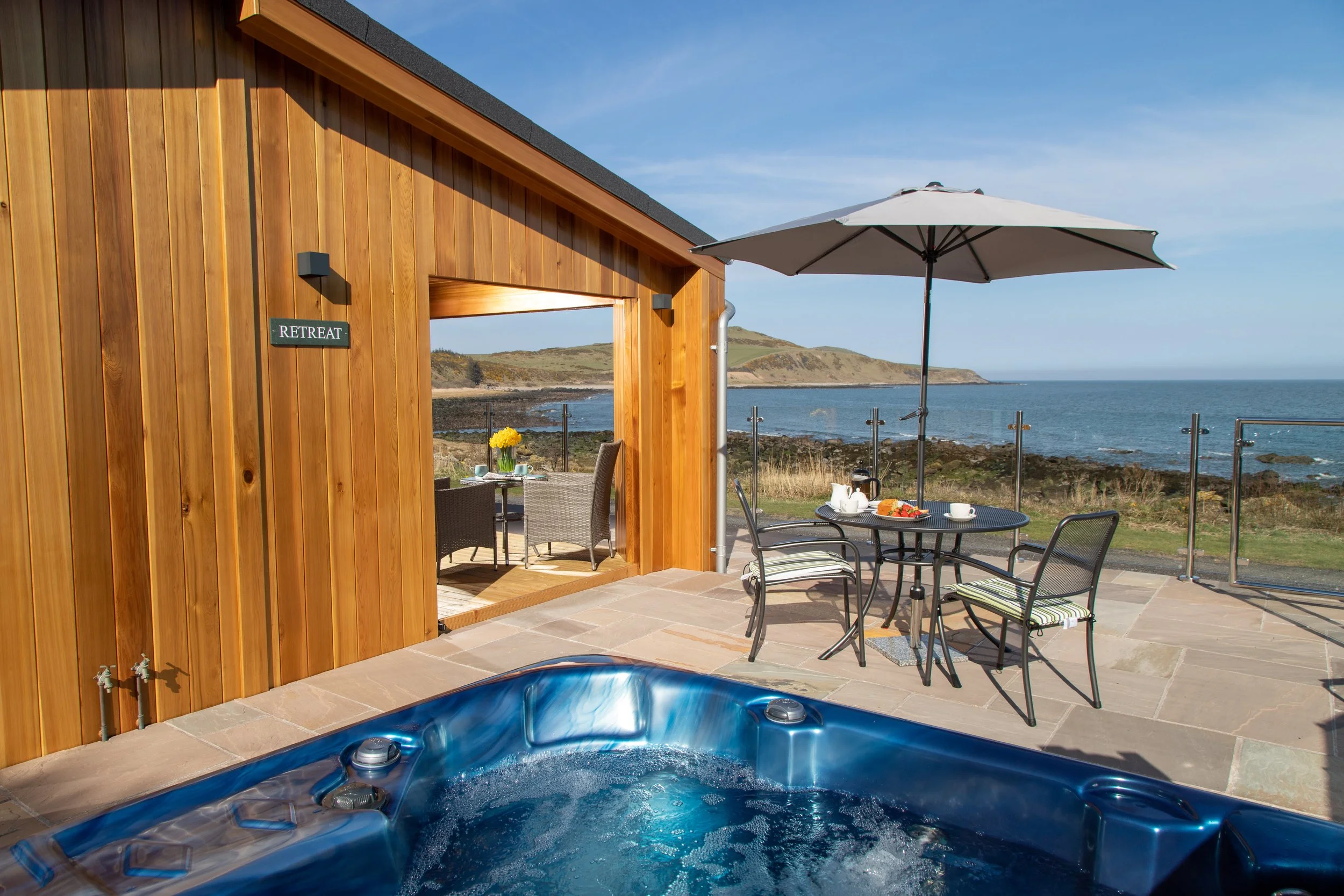 Outdoor patio with a hot tub, dining table with an umbrella, and a scenic ocean view with hills in the distance.