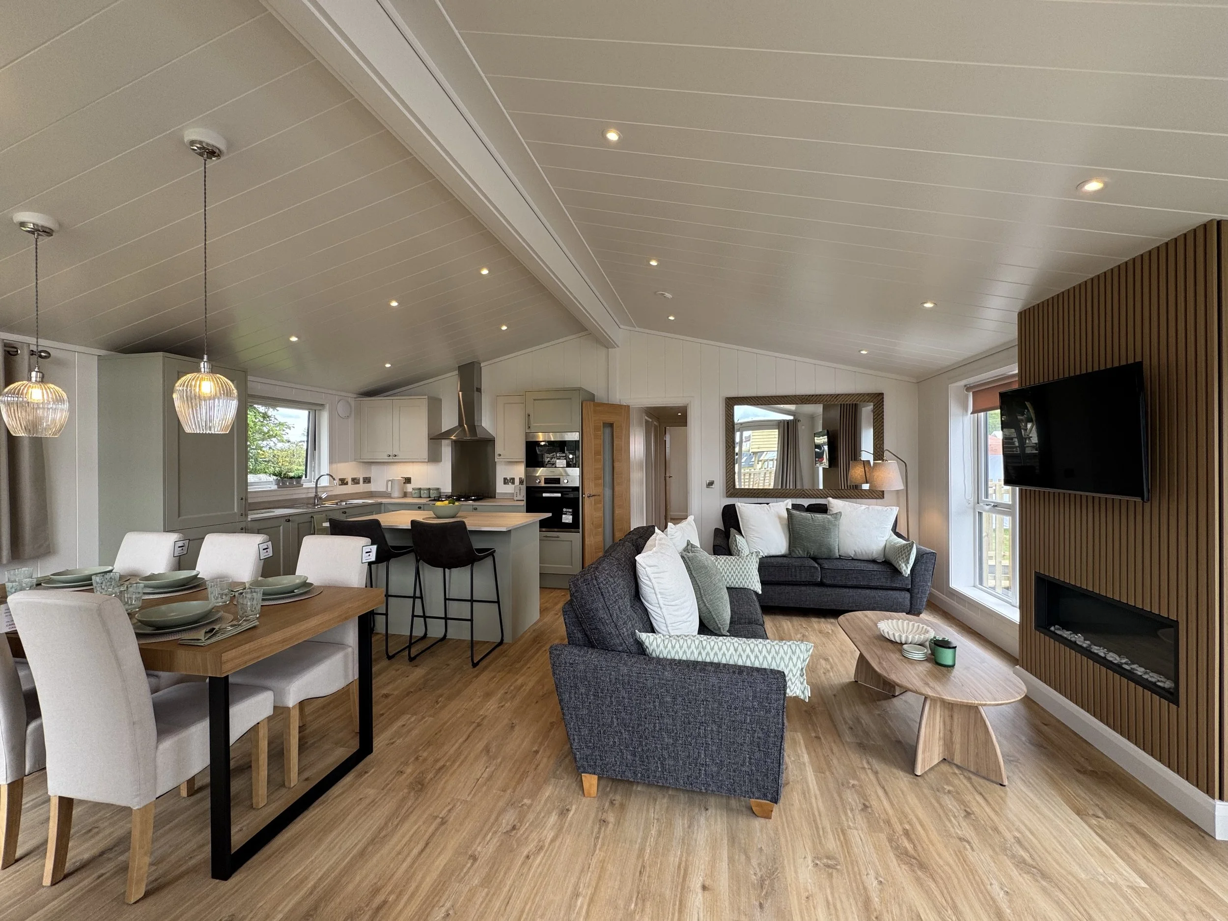 Open-concept living room and kitchen with beige and wooden tones, gray sofas with white and green pillows, wooden coffee table, dining table with white chairs, kitchen island with black barstools, and a wall-mounted TV on wood paneling.