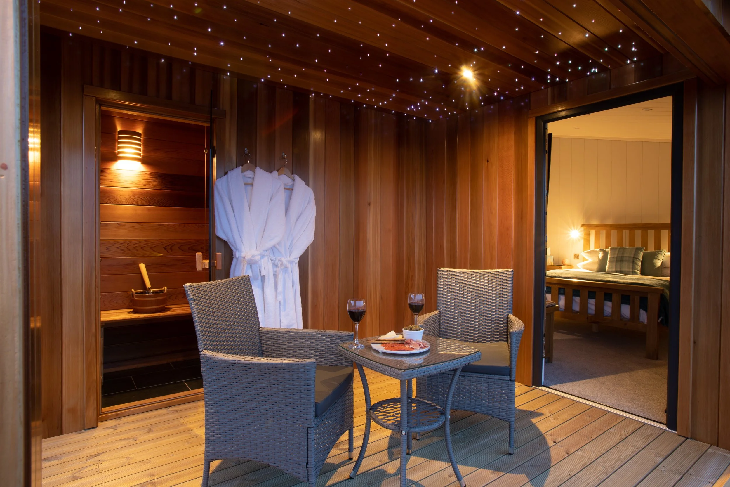 A cozy wooden porch with two wicker chairs, a round table with wine glasses, food, and a charcuterie board, adjacent to a sauna room with robes hanging, and a bedroom visible through a doorway, all illuminated warmly.