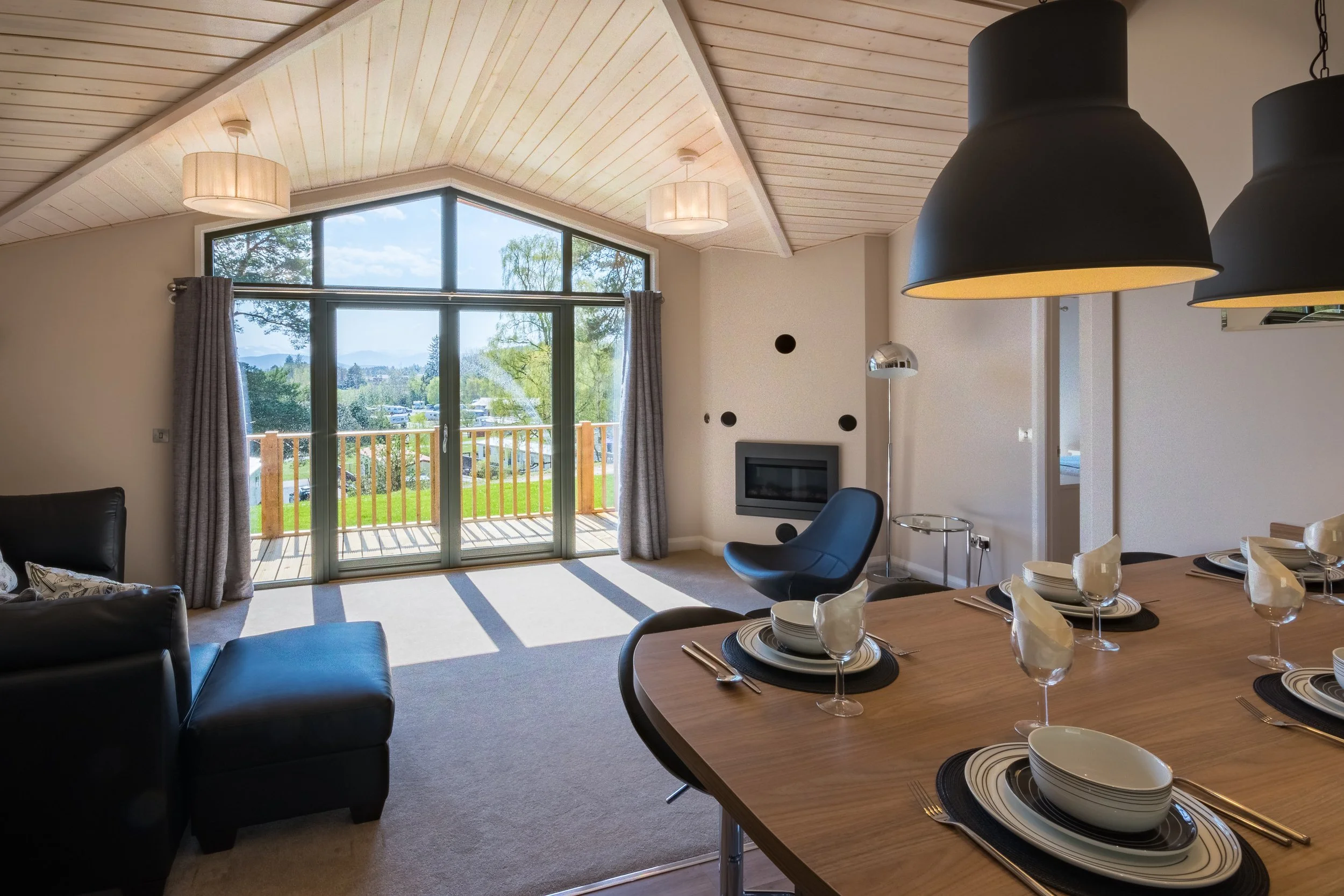Living and dining area with large window showing outdoor view, wooden ceiling, hanging lights, black leather sofa, dining table set with plates, glasses, and napkins, black and blue chairs, fireplace, and modern decor.