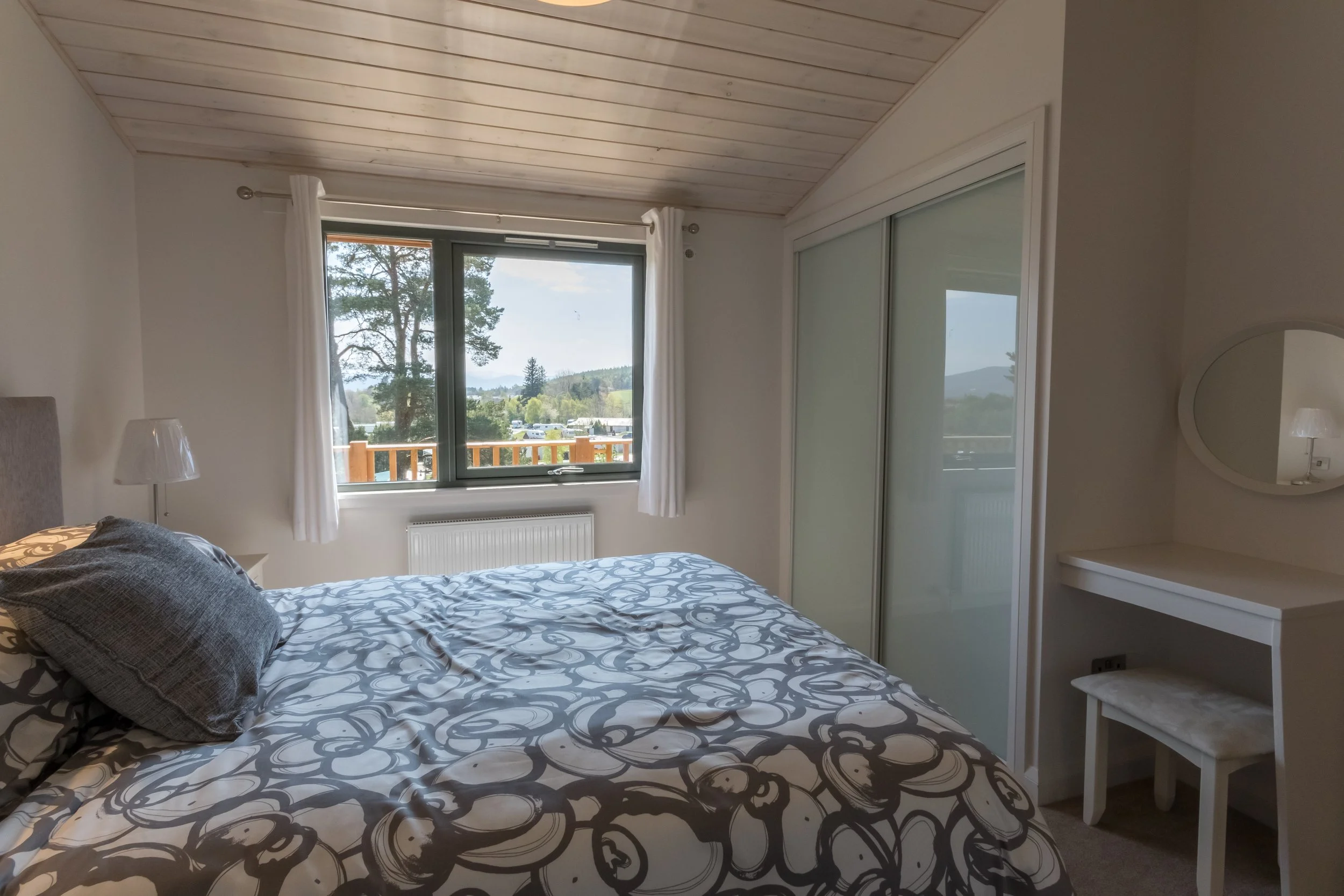 Bedroom with a bed, pillows, nightstand with lamp, window with white curtains, view of trees and mountains, built-in closet with frosted sliding doors, small white vanity with mirror and stool.