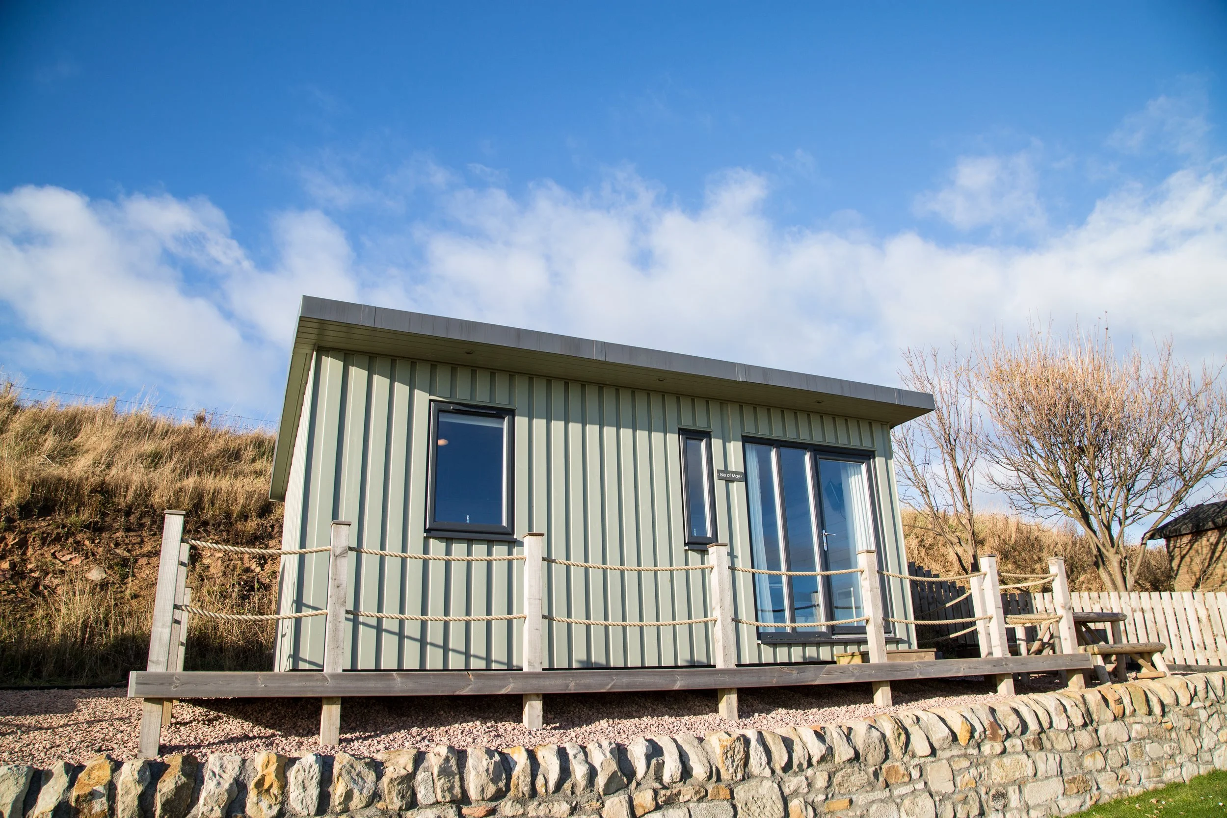 A modern tiny house with vertical green siding, large windows, and a sliding glass door, situated on a stone foundation with a small wooden deck and rope railing, surrounded by a grassy hillside and clear blue sky.