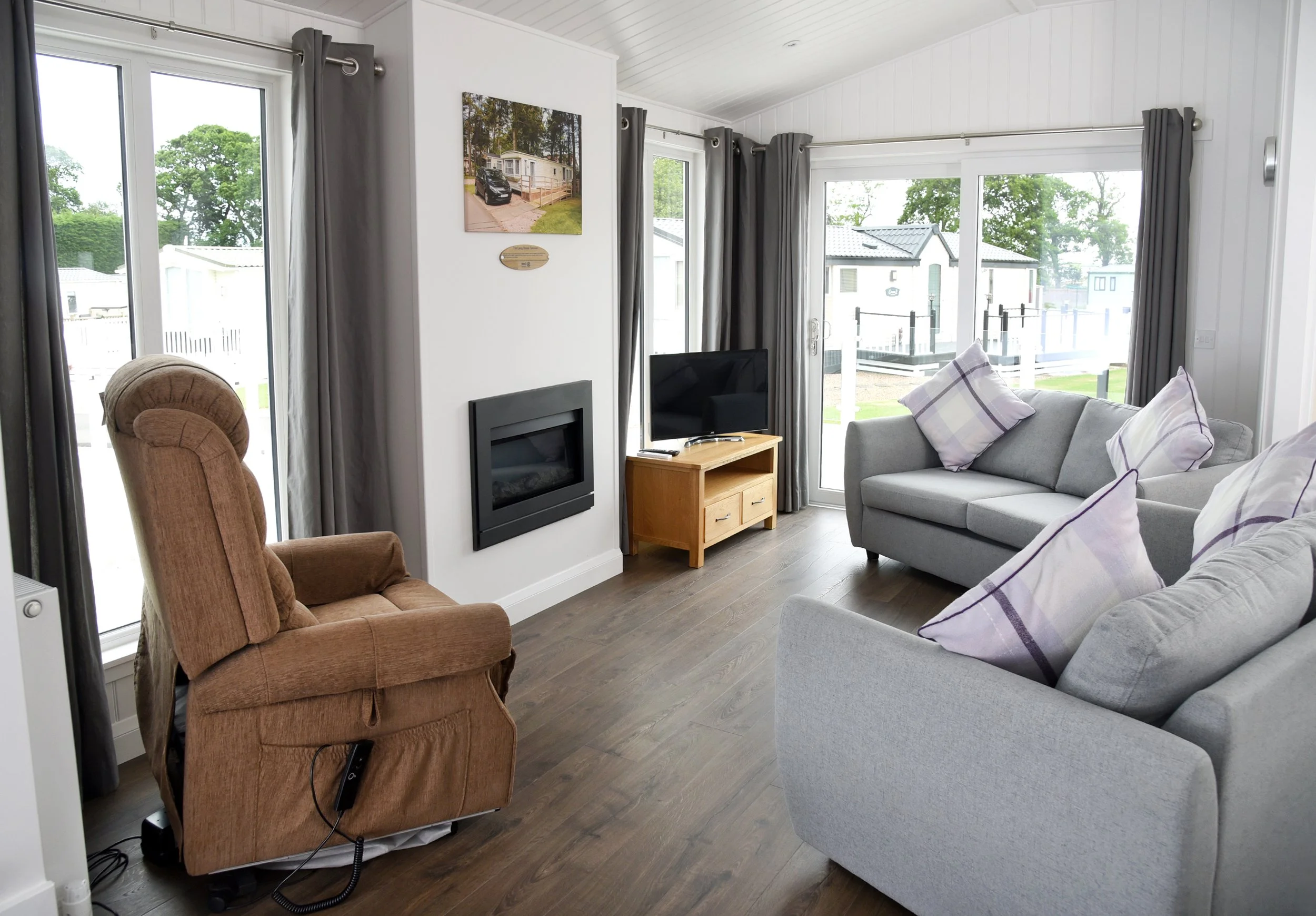 Living room with gray couches, a brown armchair, a small wooden TV stand with a flat-screen TV, a wall-mounted fireplace, large windows with gray curtains, and a view of neighboring mobile homes outside.
