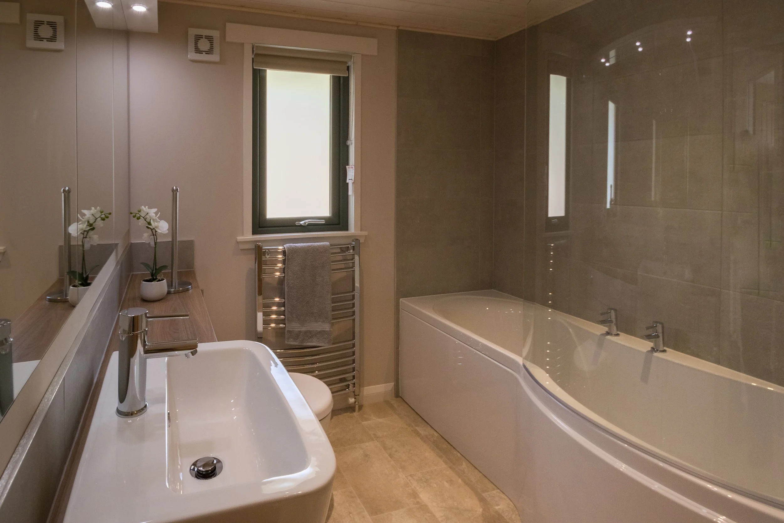 Modern bathroom with a white bathtub, a sink with a chrome faucet, a small window, and a heated towel rail with a gray towel.