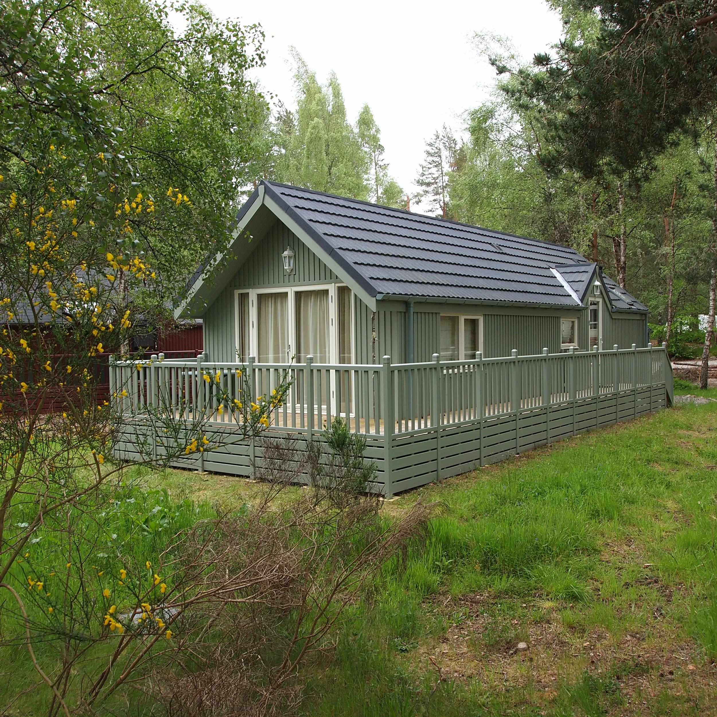 A gray house with a large wrap-around deck, situated in a green, wooded area with trees and grass.