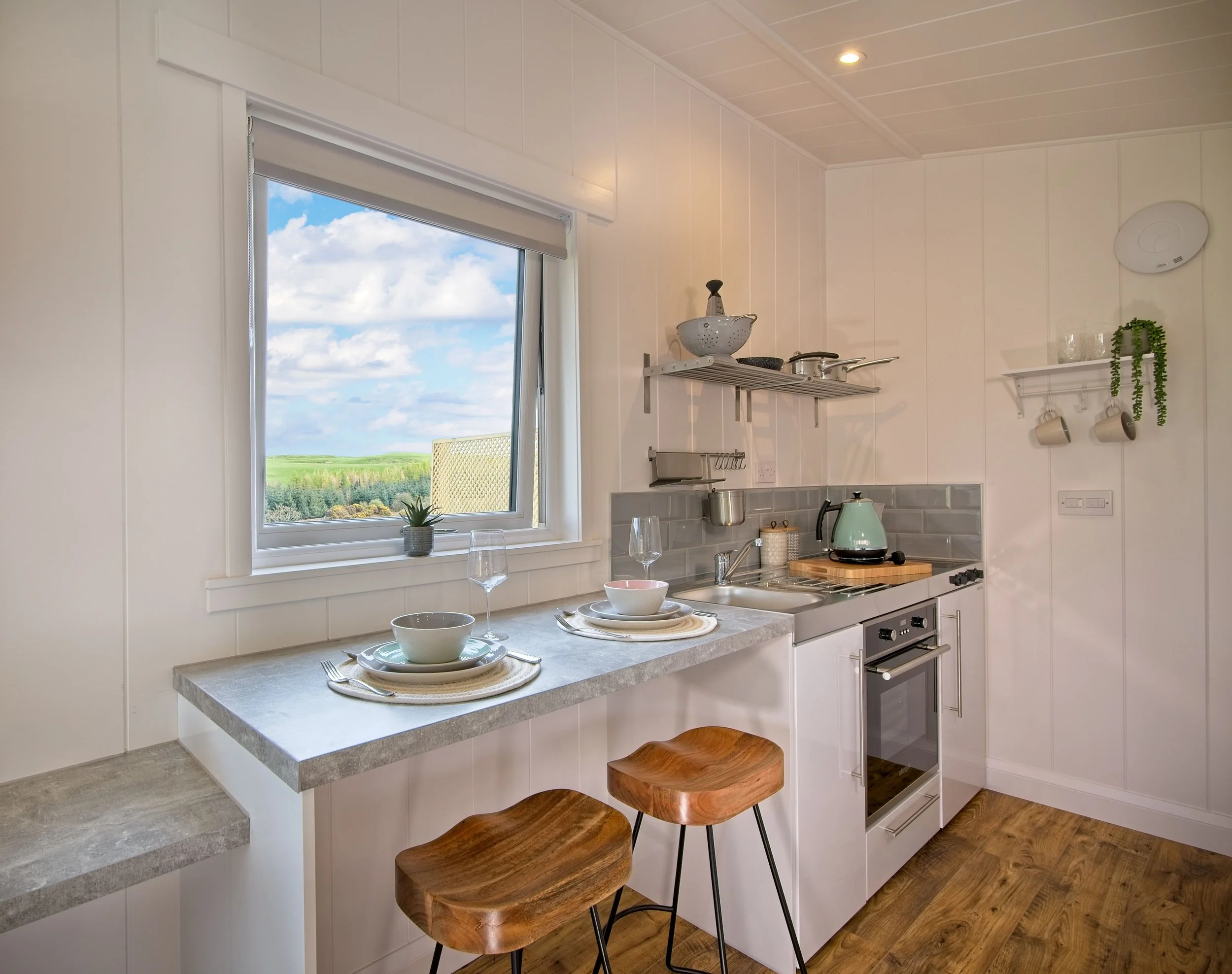 Cozy kitchen with white walls, a large window showing a green landscape and blue sky, a small counter with bowls and plates, a stove with a kettle, shelves with kitchenware, and wooden bar stools.