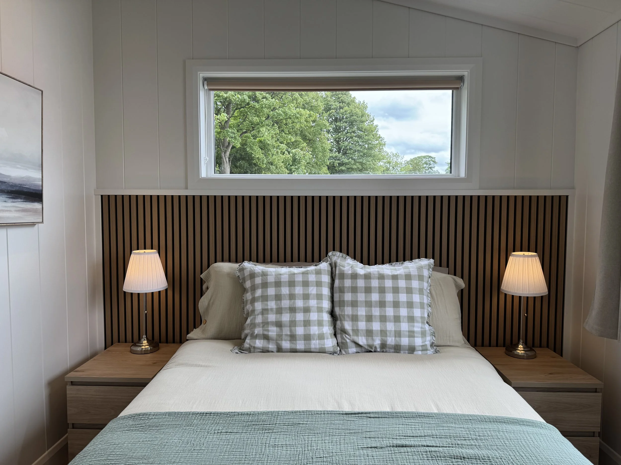 A bedroom with a bed, pillows, side tables with lamps, a window with trees outside, and artwork on the wall.