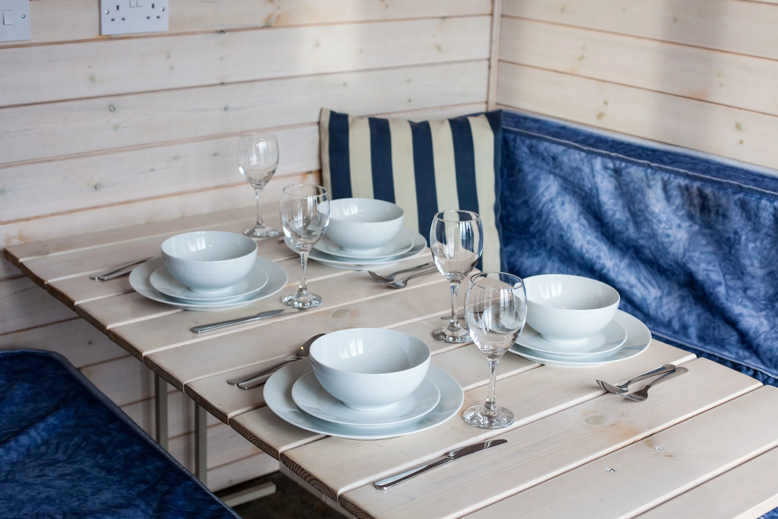 Set of white bowls, plates, wine glasses, forks, and spoons on a wooden table with blue cushioned seating and a striped pillow in a light wood-paneled room.