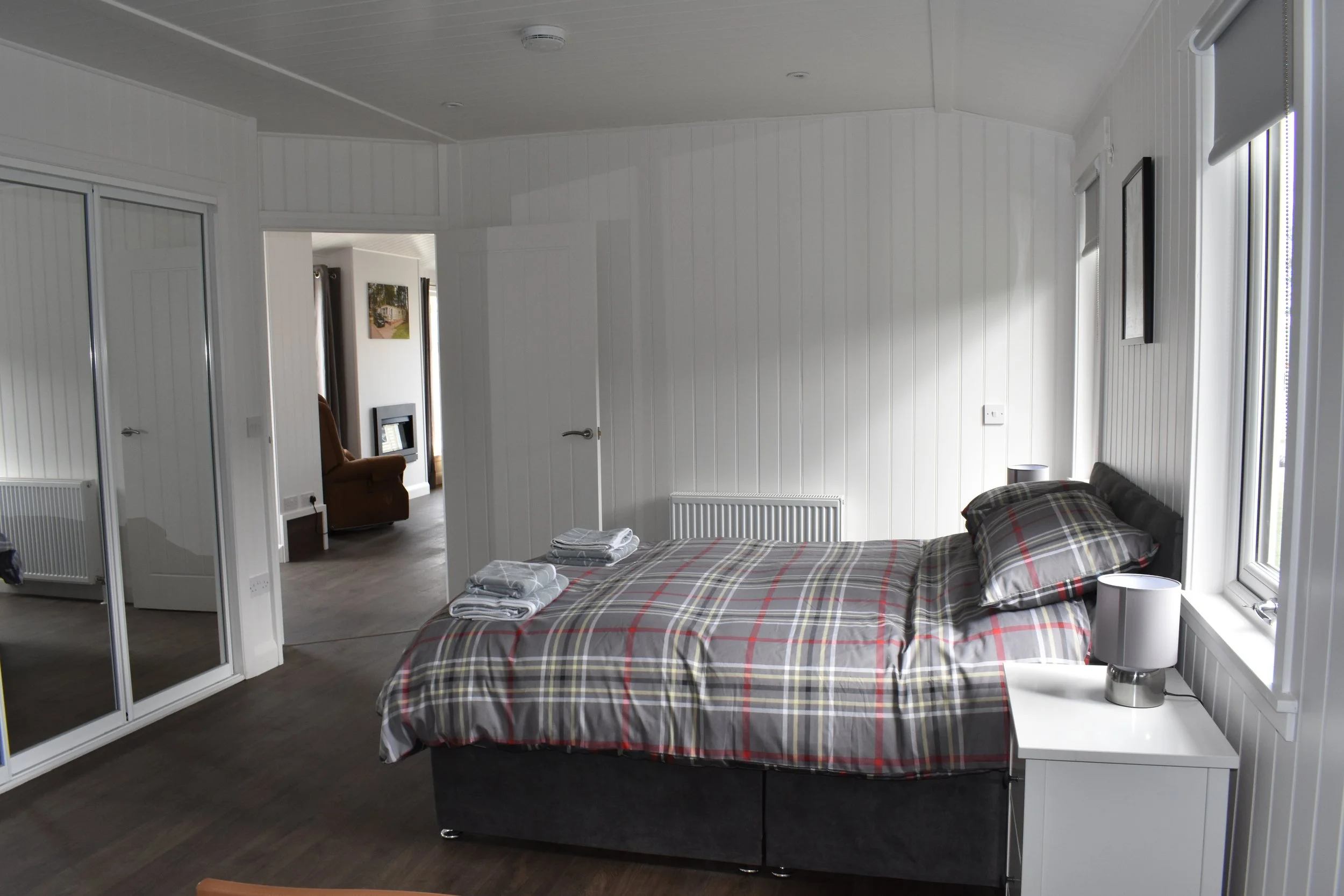 A bedroom with a bed covered in plaid bedding, two folded towels on top, white walls, a window with blinds, a small white nightstand with a lamp, and a mirror closet door.