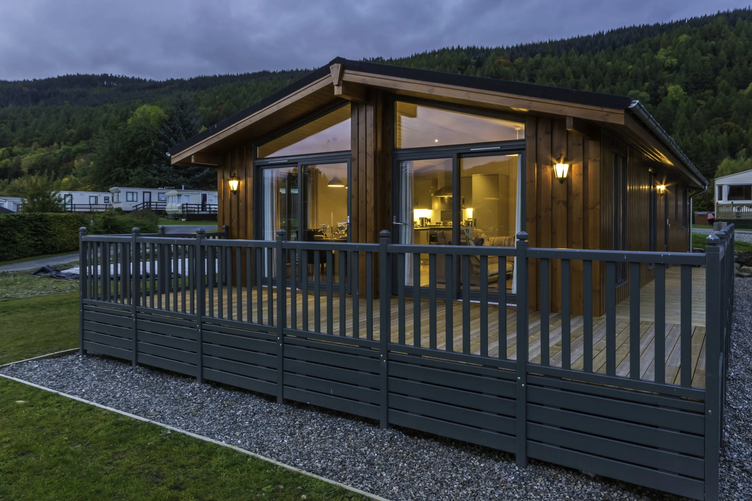 A modern wooden house with large glass sliding doors, illuminated with warm lights, surrounded by a fenced wooden deck, with a backdrop of green hills and trees.