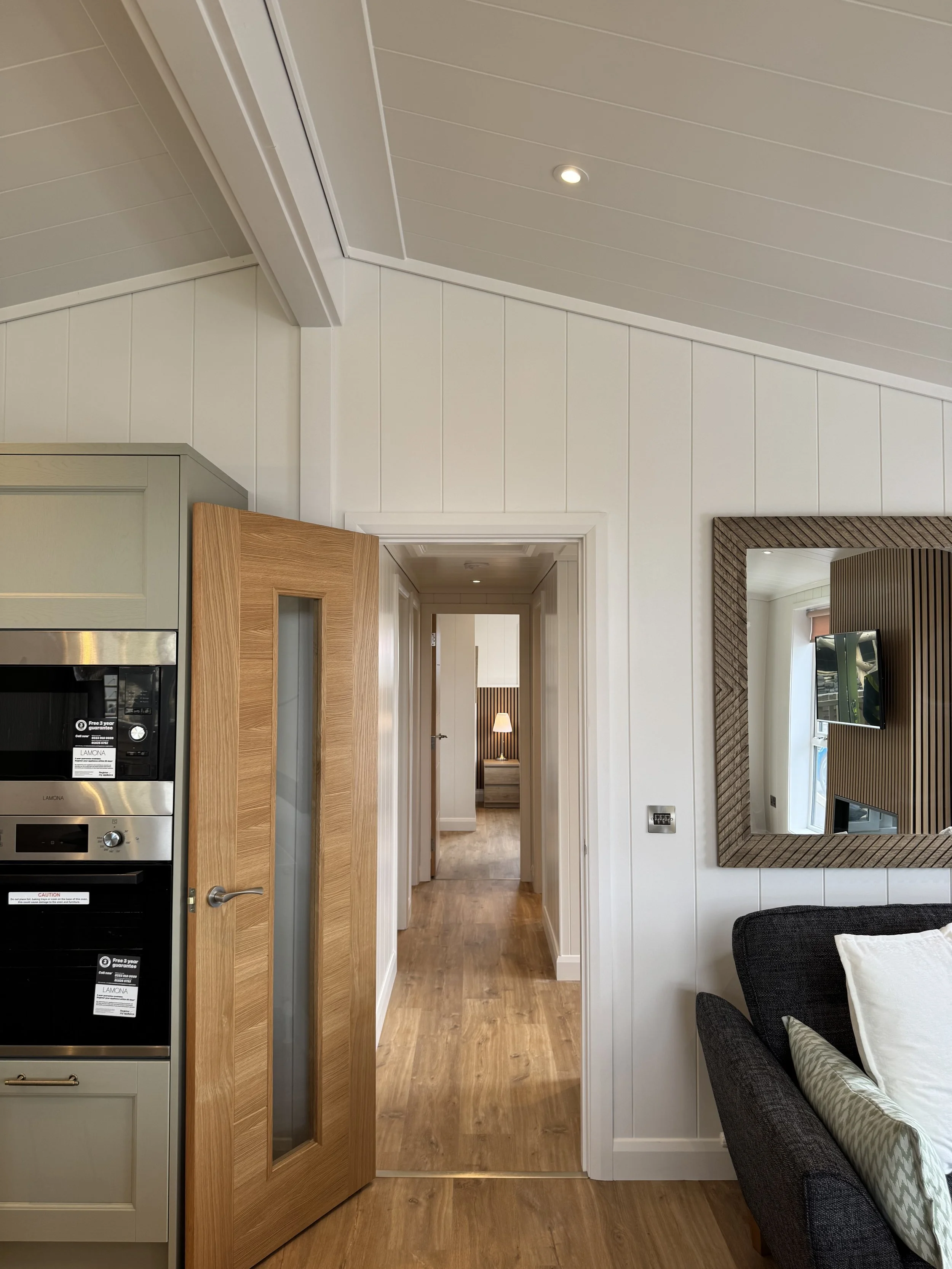 Interior view of a modern apartment with wooden flooring, white paneled walls, a door slightly open leading to a hallway, and a mirror on the wall reflecting part of a living room.