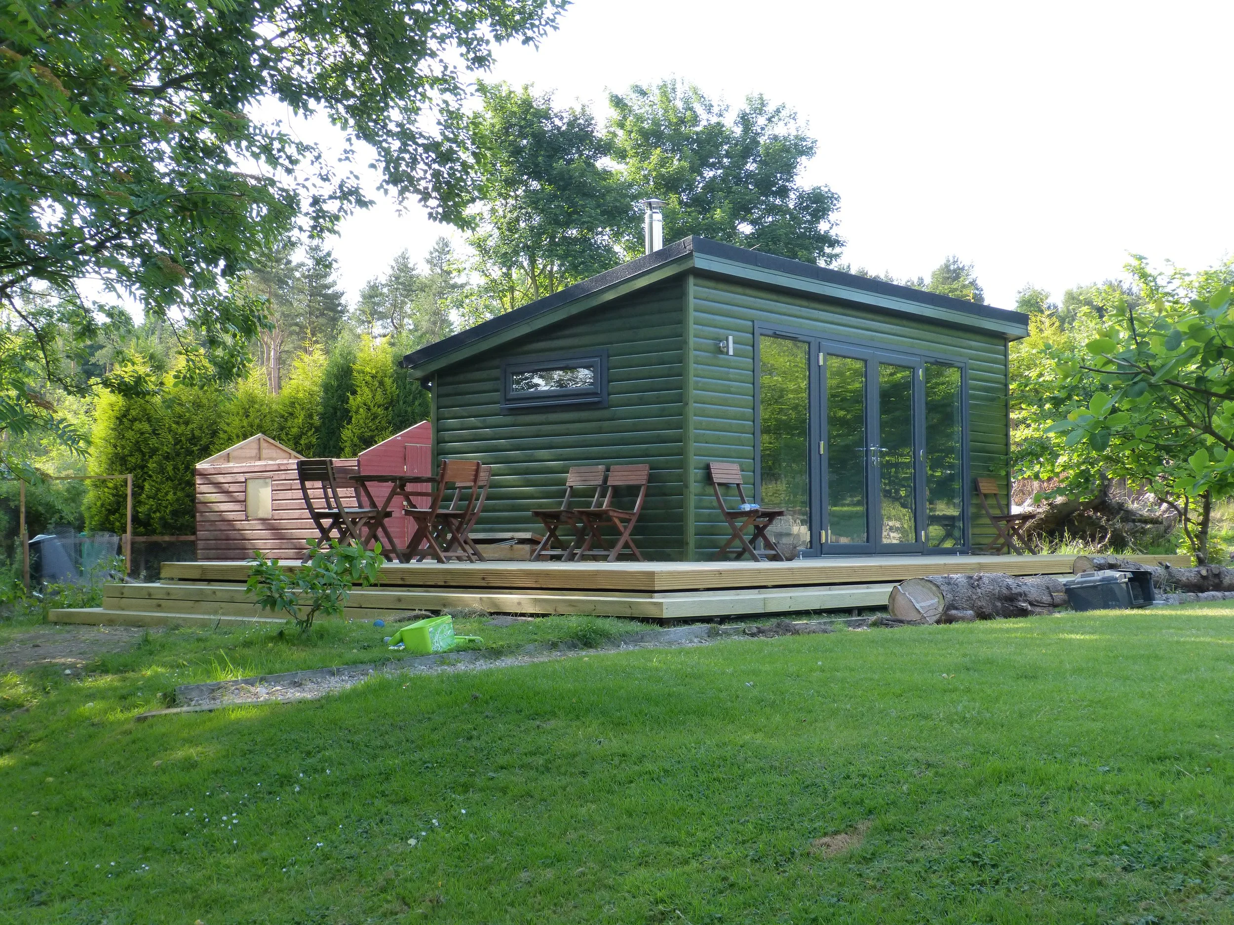 Green tiny house with large glass doors and windows, wooden deck with outdoor furniture, surrounded by lush green trees and lawn.