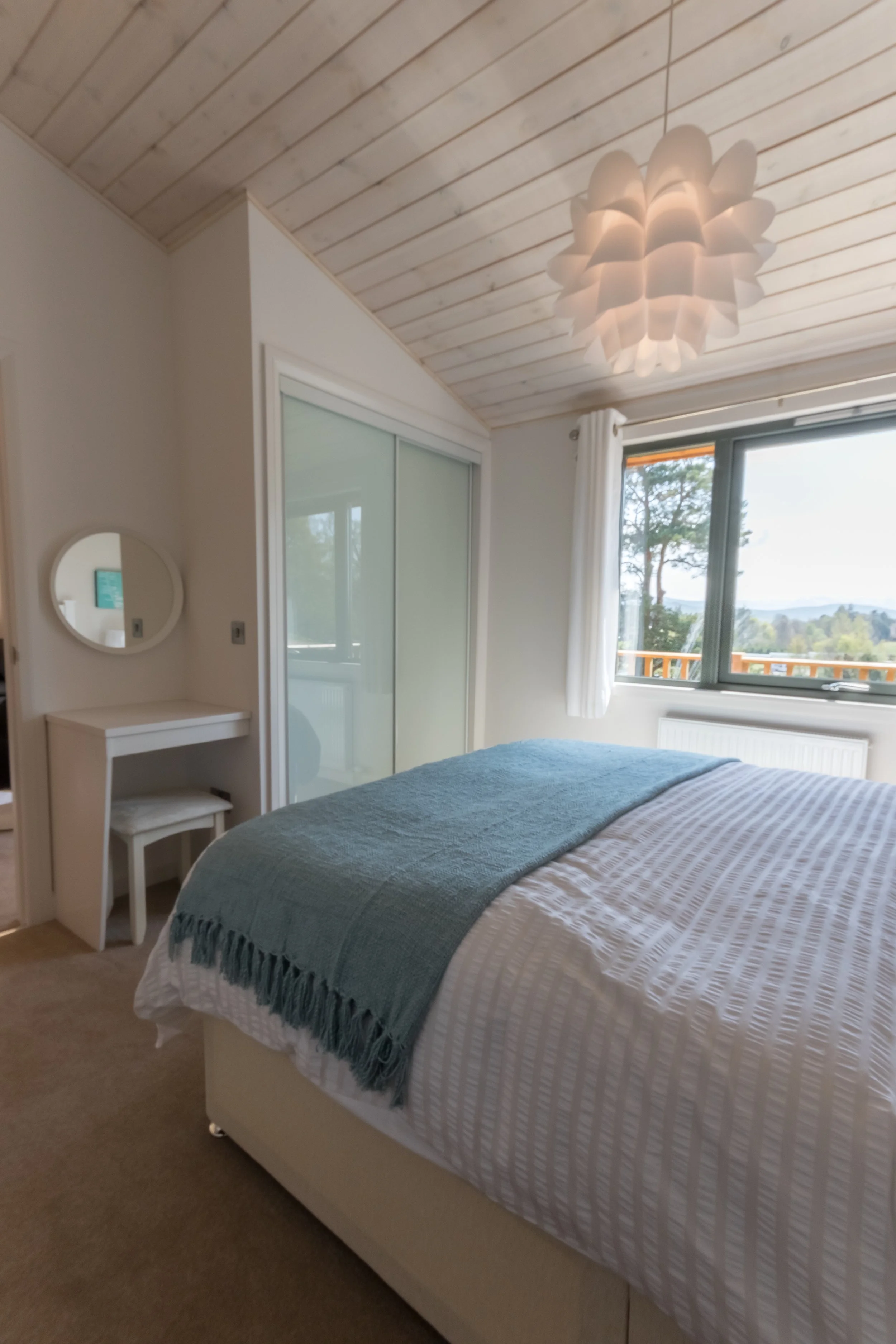 A cozy bedroom featuring a bed with a white striped duvet and a teal throw blanket, a window with a view of trees, a small vanity with a round mirror, and a modern white ceiling light fixture.