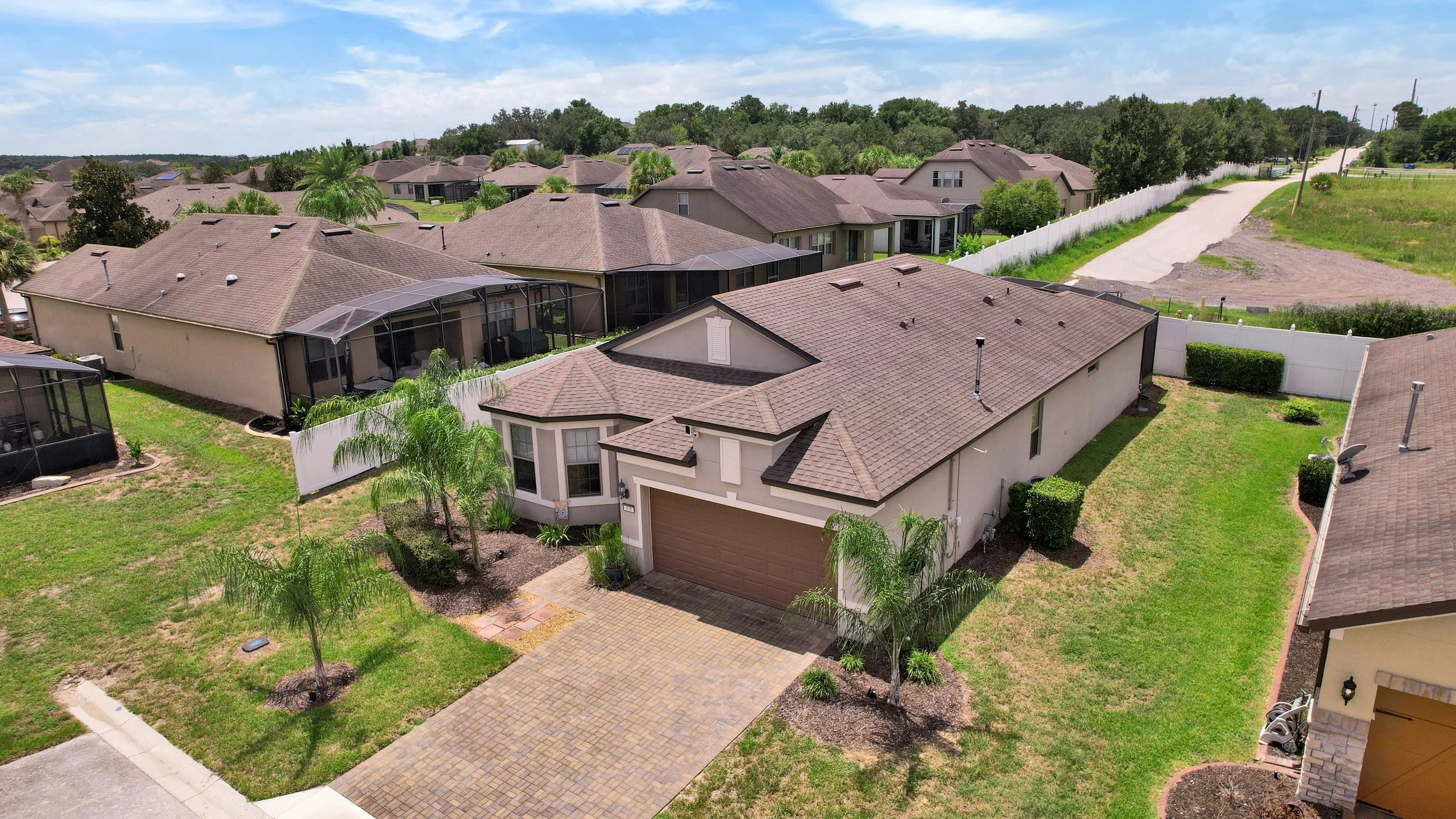 Aerial view of a suburban neighborhood showing multiple houses with brown tiled roofs, green lawns, palm trees, and a white fence along a dirt road on a partly cloudy day.
