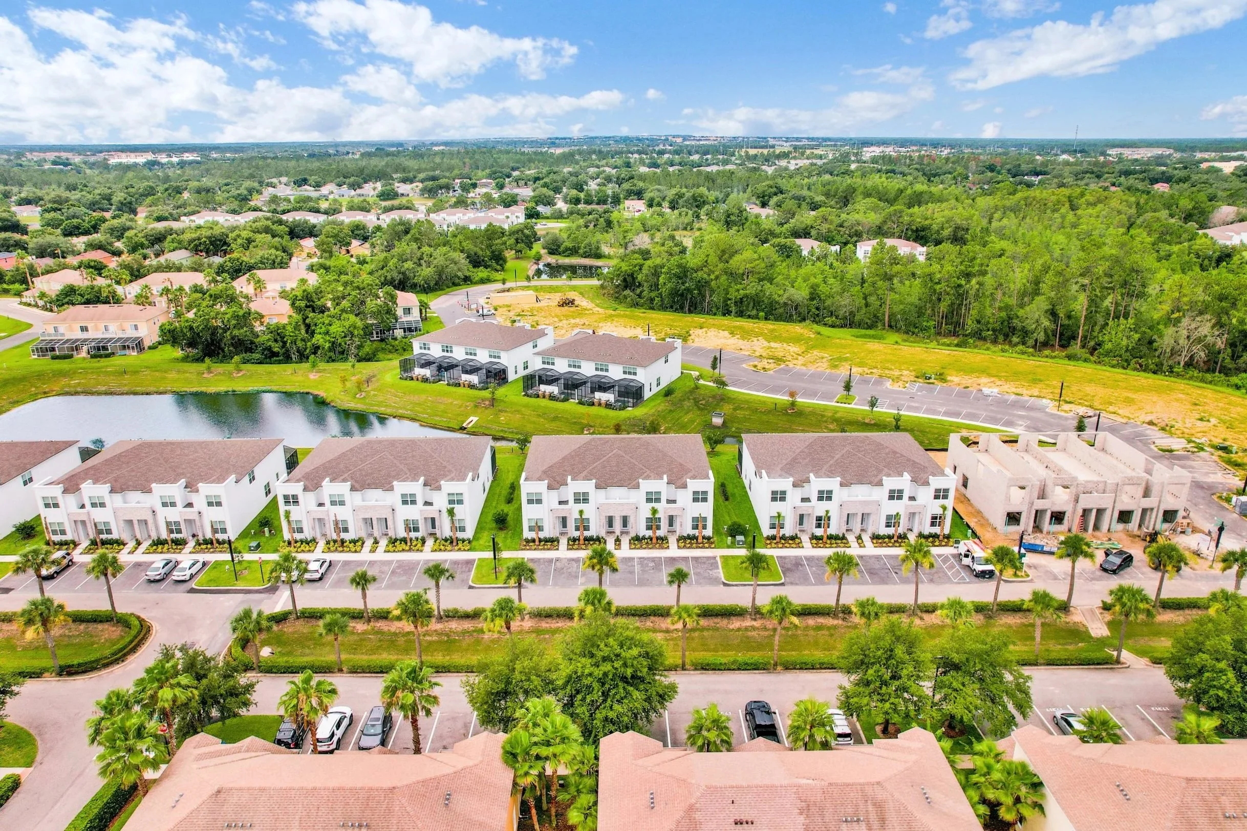 Aerial view of a residential area with white townhouses, a pond, and green trees, with parking lots and a wooded area in the background.