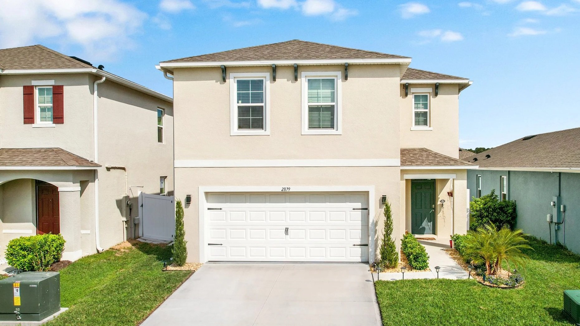 Front view of a beige two-story house with white garage door, green front door, and small landscaped yard. There are neighboring houses on either side, with a 