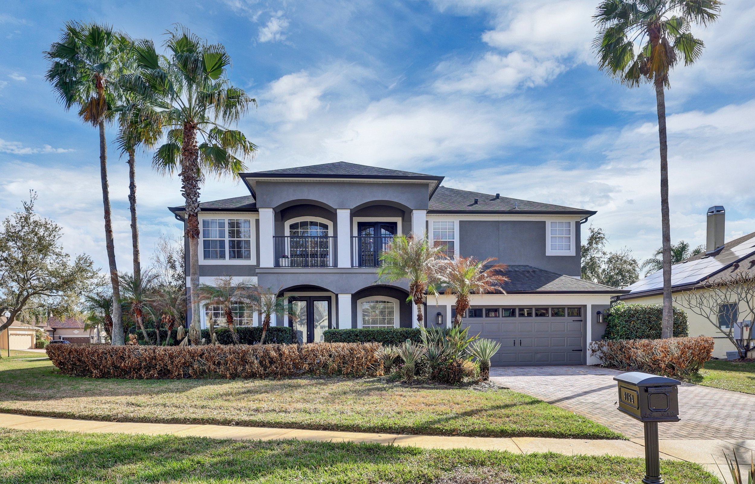 Front of a house with a green garage door, two bushes, potted plants, and a satellite dish on the roof under a cloudy sky.