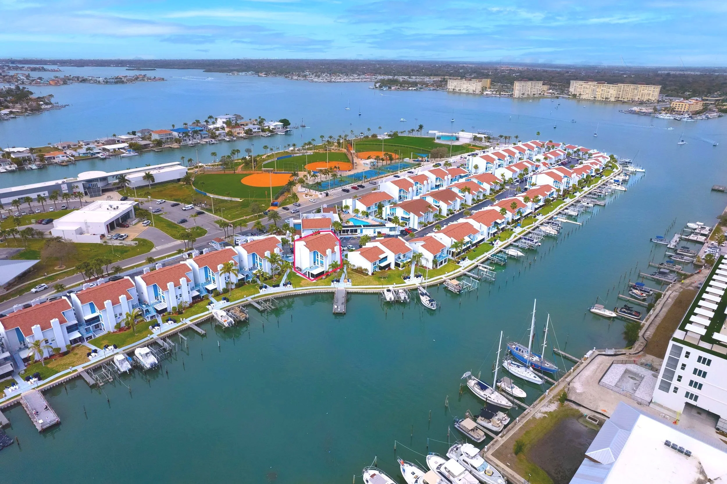 Aerial view of waterfront residential buildings with boats docked along the marina, a baseball field, and water in the background.